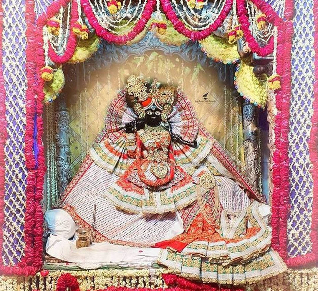krishna god 桌布 壁紙 — 一尊裝飾華麗的黑皮膚印度教女神雕像，在紅、黃、綠色花環組成的鮮豔花冠下威嚴地坐著。