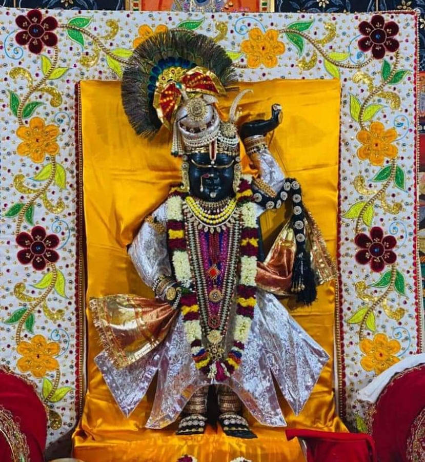 Shrinathji 桌布 壁紙 — 一尊裝飾華麗的黑色神像，頭戴孔雀羽毛冠冕，佩戴黃金珠寶，坐在金色台座上，周圍環繞著色彩繽紛的花卉刺繡。