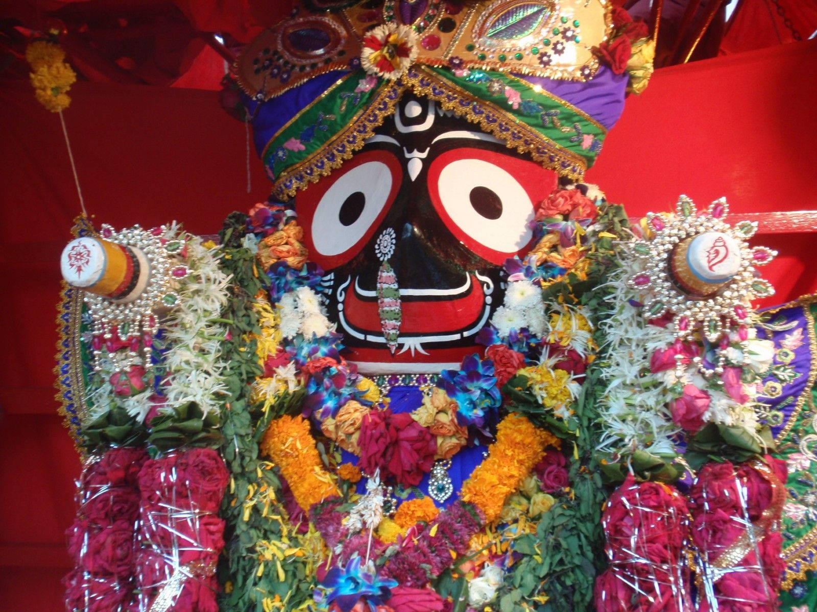 Jagannath 桌布 壁紙 — 一尊裝飾鮮豔的印度教神像，以彩色花朵和珠寶點綴，背景為紅色。