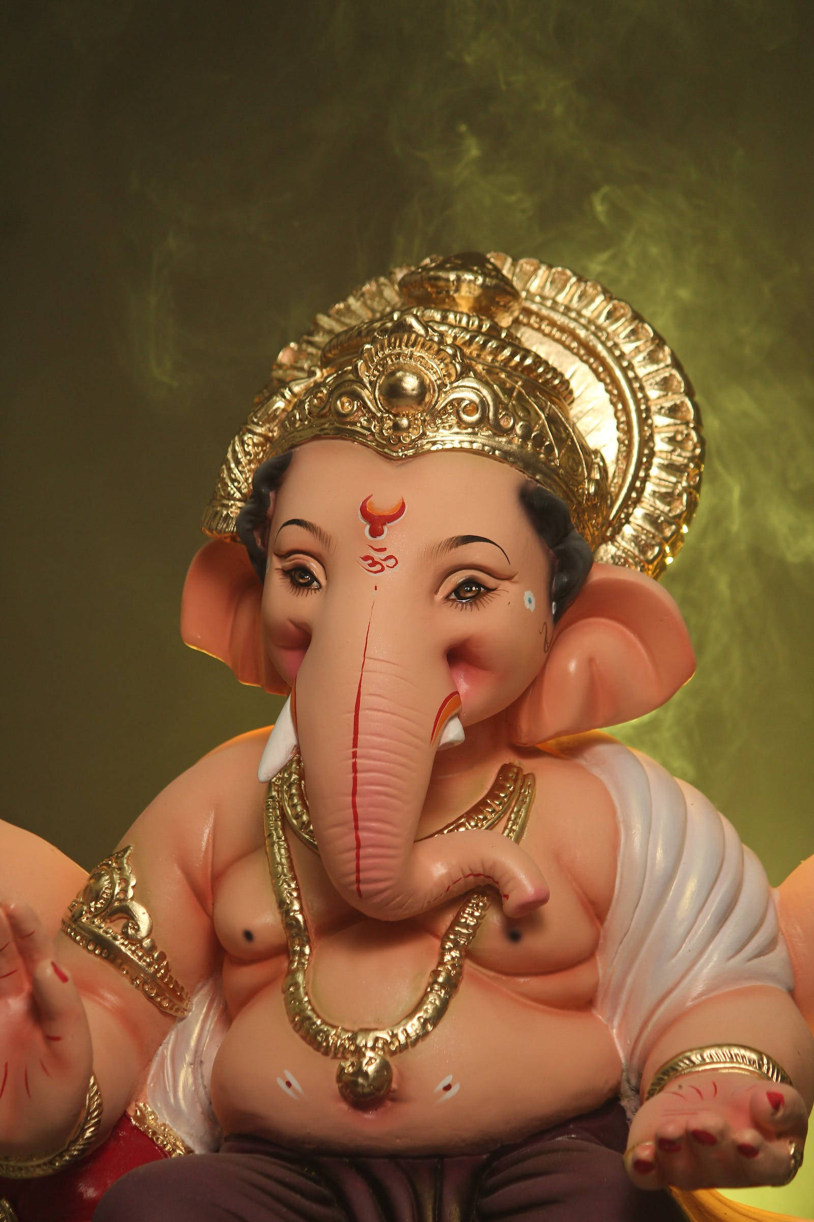 Vinayaka 桌布 壁紙 — 一尊詳細的象頭神像，飾有黃金首飾和皇冠，背後煙霧繚繞，襯托著溫暖的綠色背景。