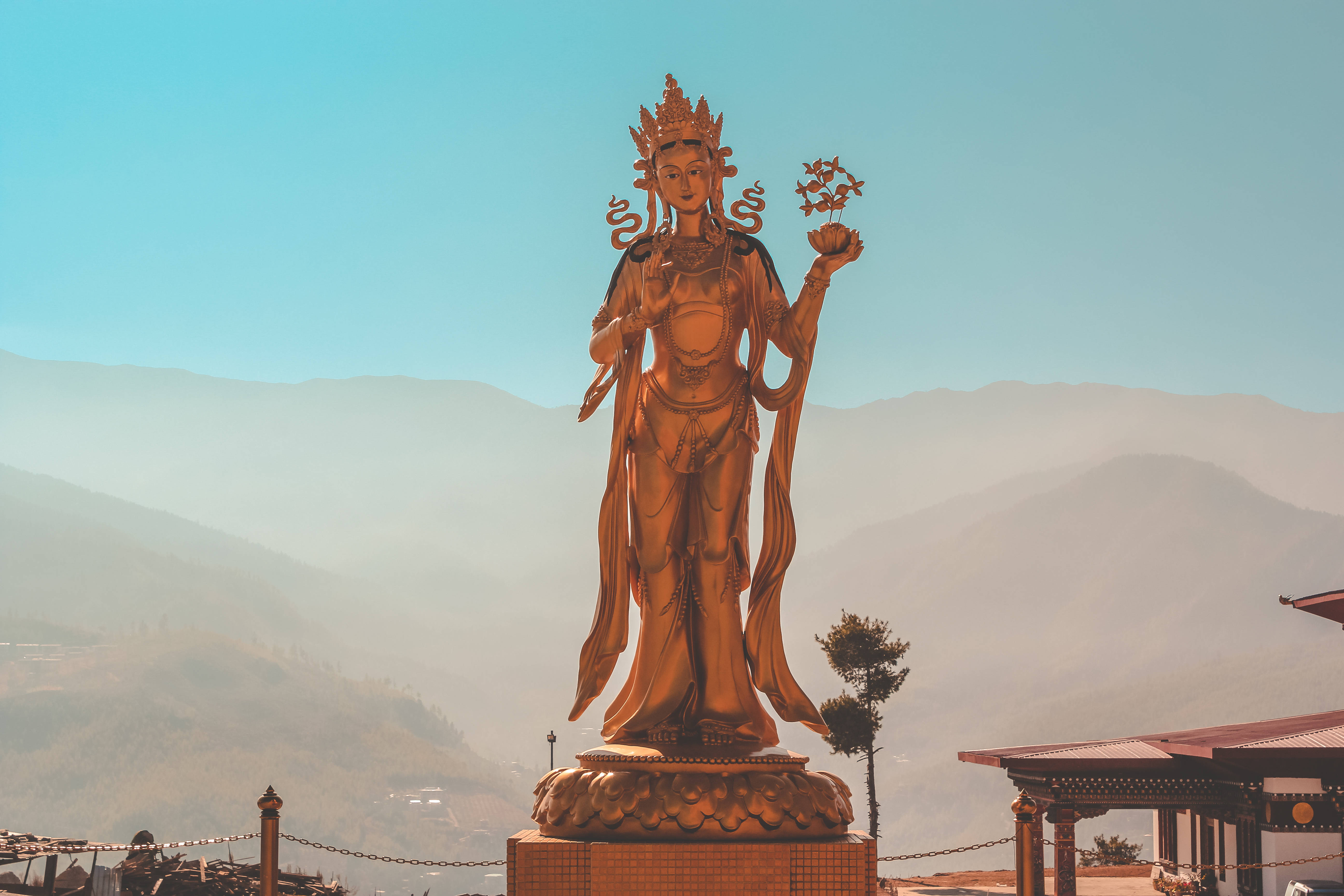 Bhutan 桌布 壁紙 — 一尊金色佛像安詳地立於基座上，背景是晴朗藍天下朦朧的山巒。
