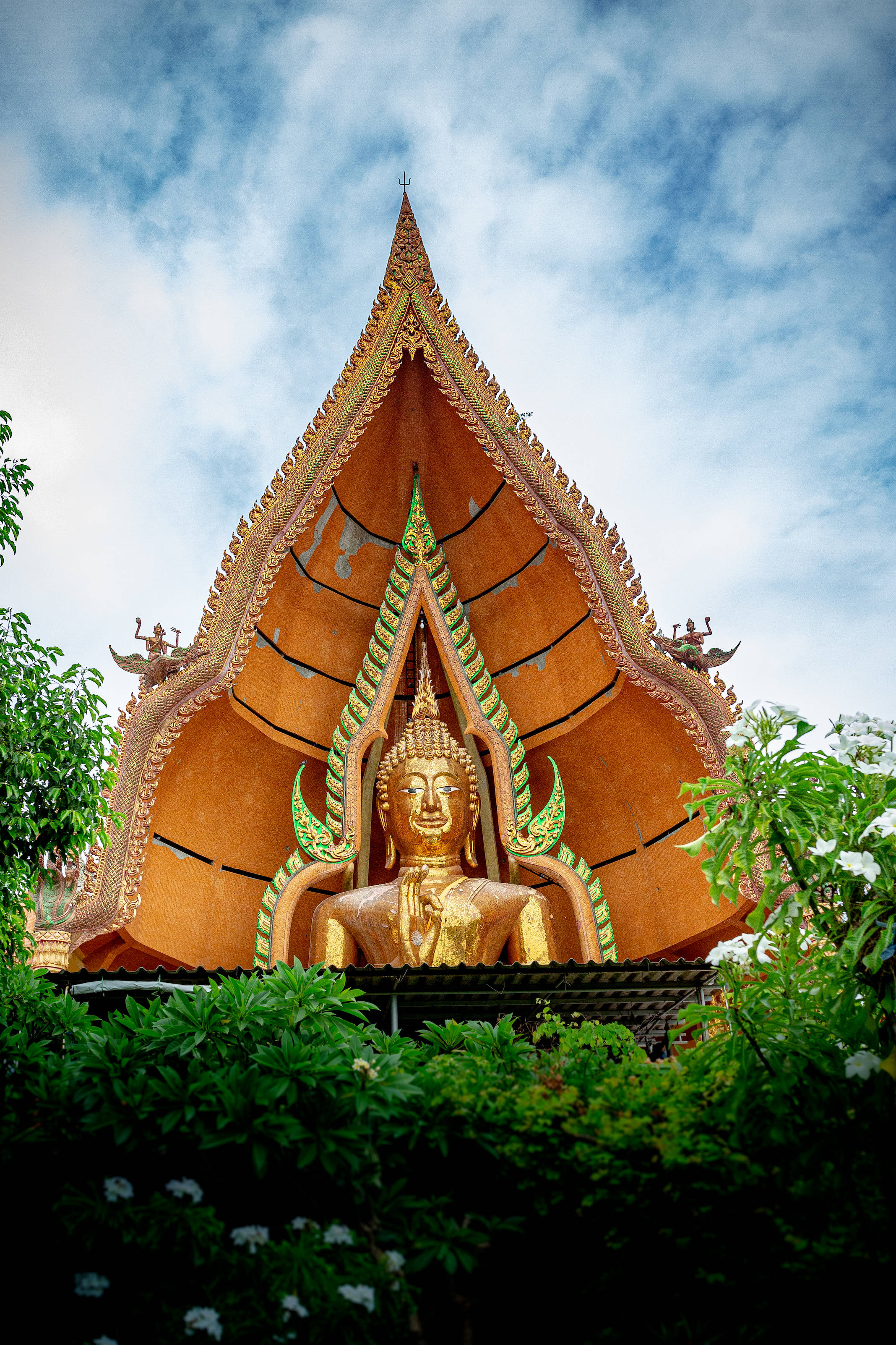 Gautam Buddha 桌布 壁紙 — 一尊金色佛像寧靜地坐在裝飾著綠色點綴的鮮橙色寺廟屋頂下，周圍環繞著茂密的綠色植物，天空部分多雲。