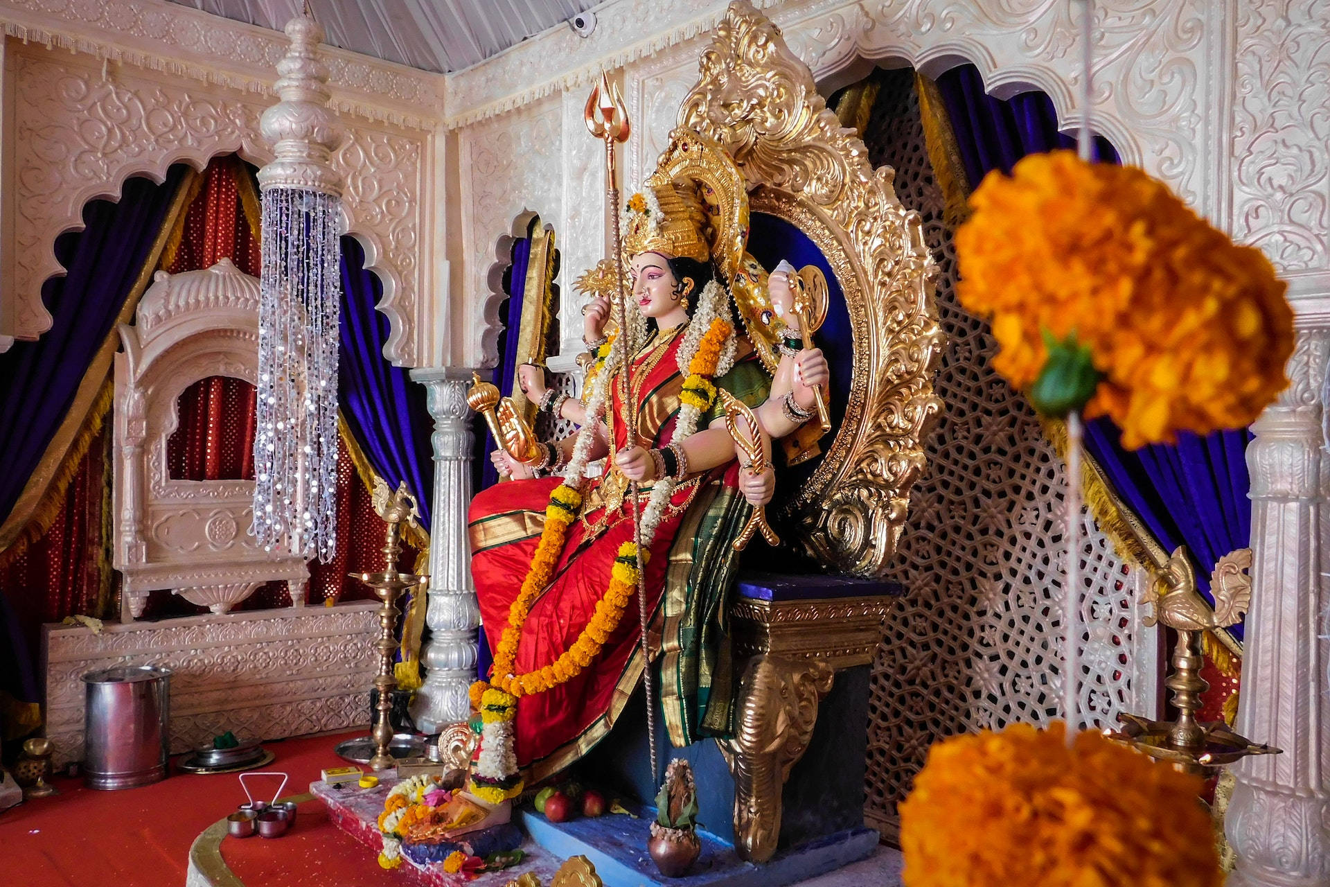 Hindu Temple 桌布 壁紙 — 一尊金色印度教女神雕像，飾以紅金兩色，莊嚴地坐在華麗的寶座上，周圍環繞著萬壽菊和精緻的寺廟裝飾。