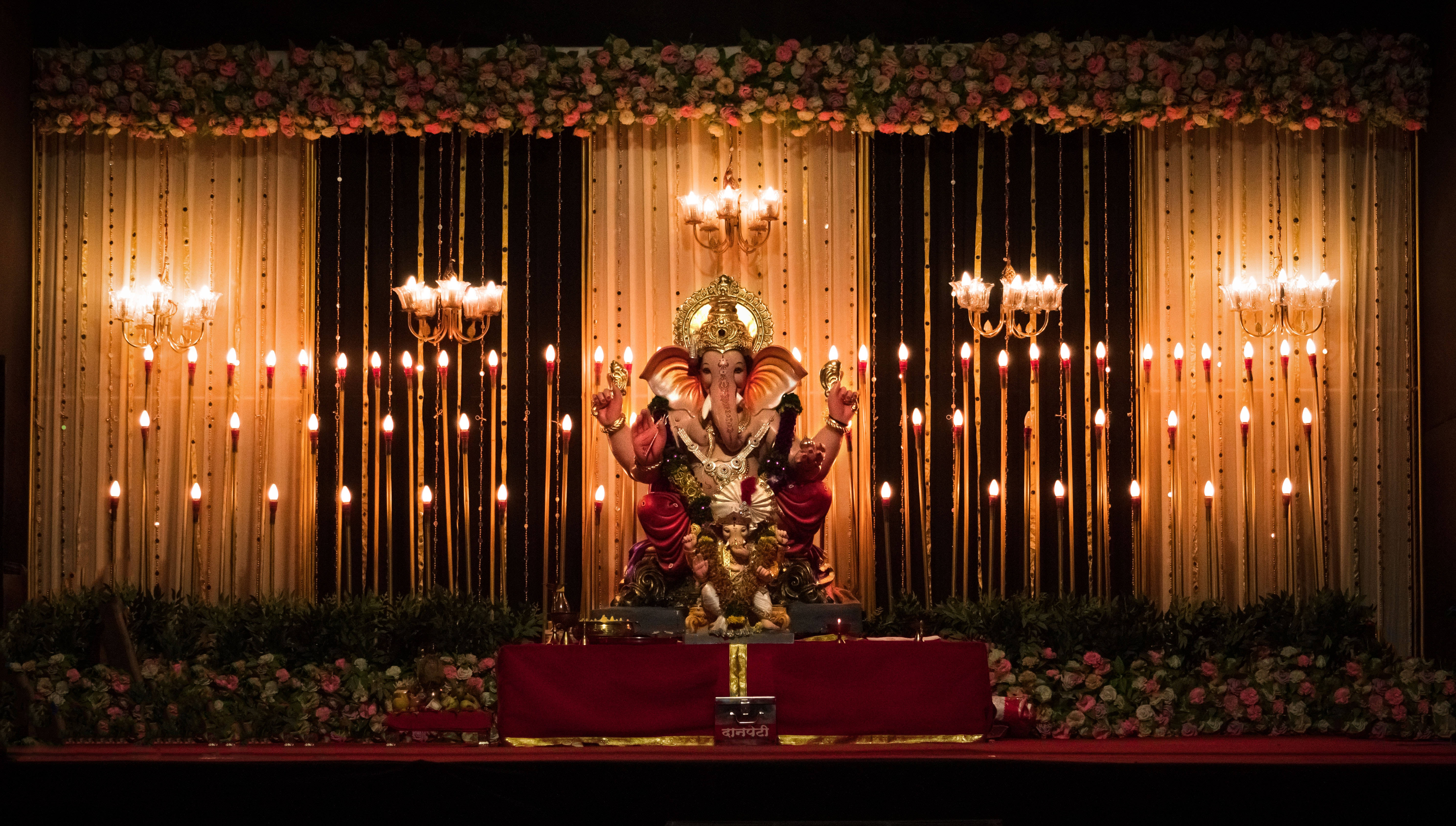 Ganesh 桌布 壁紙 — 一尊金色的象神雕像威嚴地坐在紅色高台上，周圍環繞著發光的蠟燭和花環，下方是溫暖的吊燈。