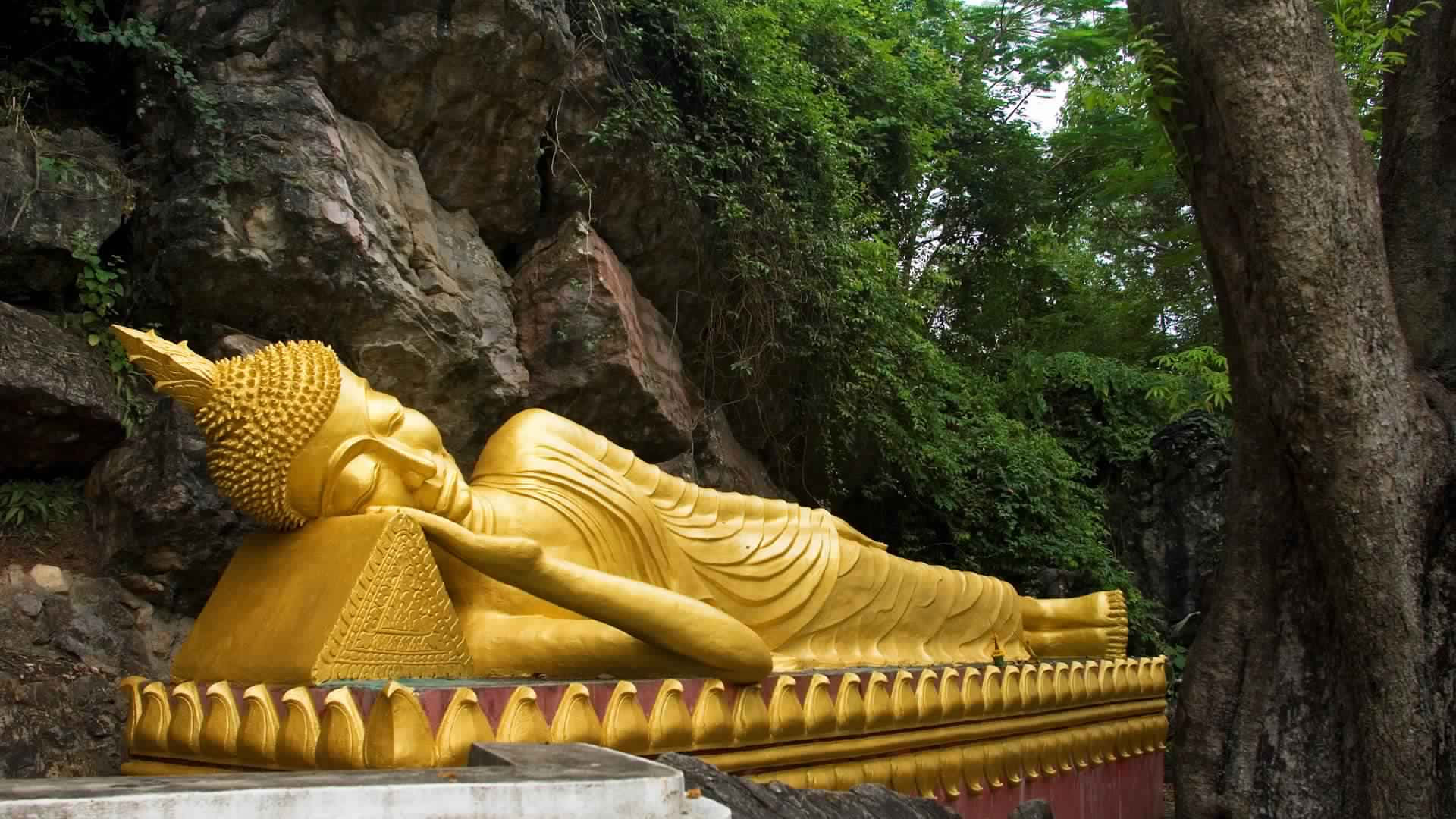 高清 桌布 壁紙 — 一尊金色臥佛安詳地躺在裝飾底座上，四周環繞著茂密的綠樹和岩石地形。
