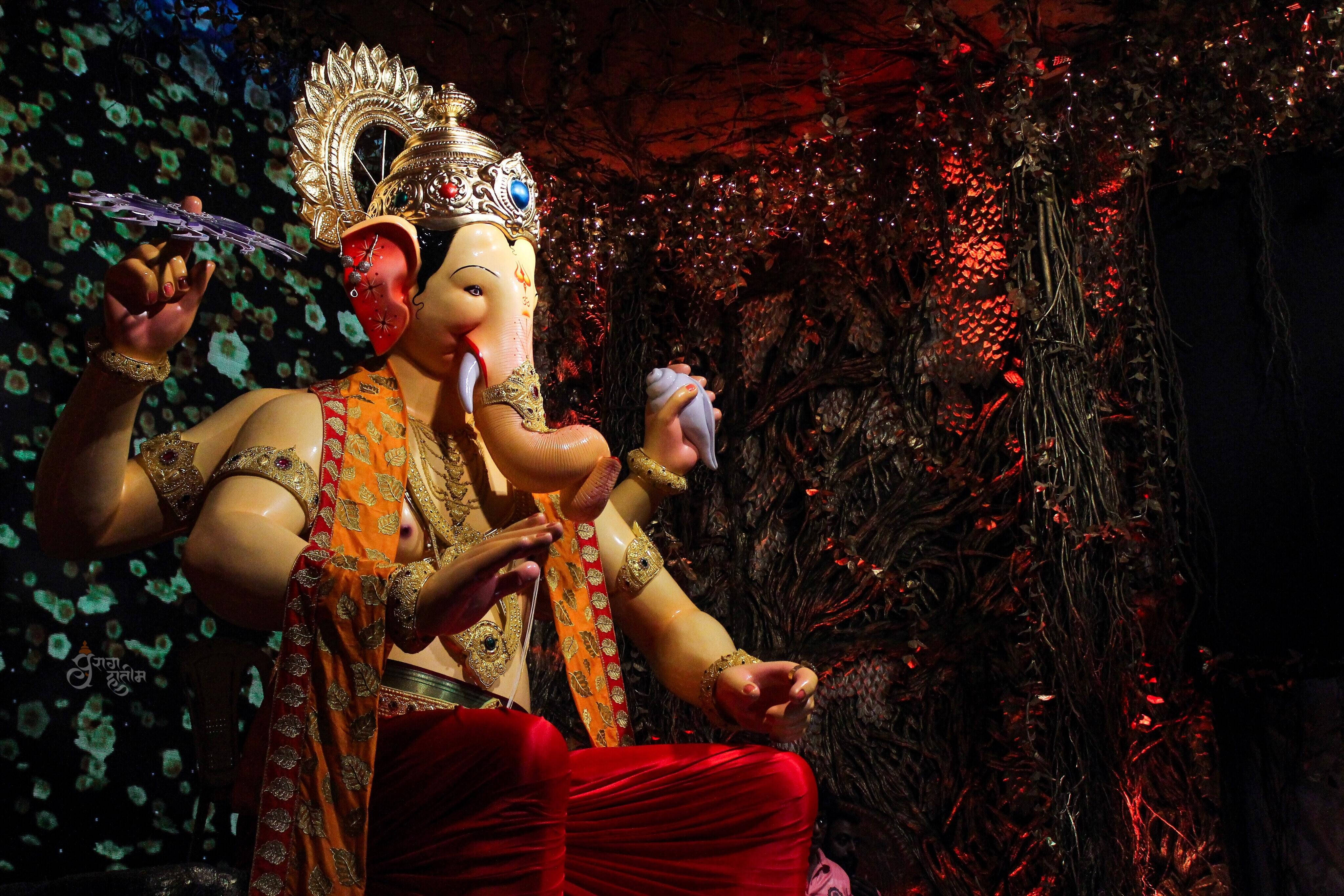ganesh 4K 桌布 壁紙 — 一尊金色象神雕像，裝飾著橙色和紅色，在深色花卉背景和戲劇性燈光下寧靜地坐著。