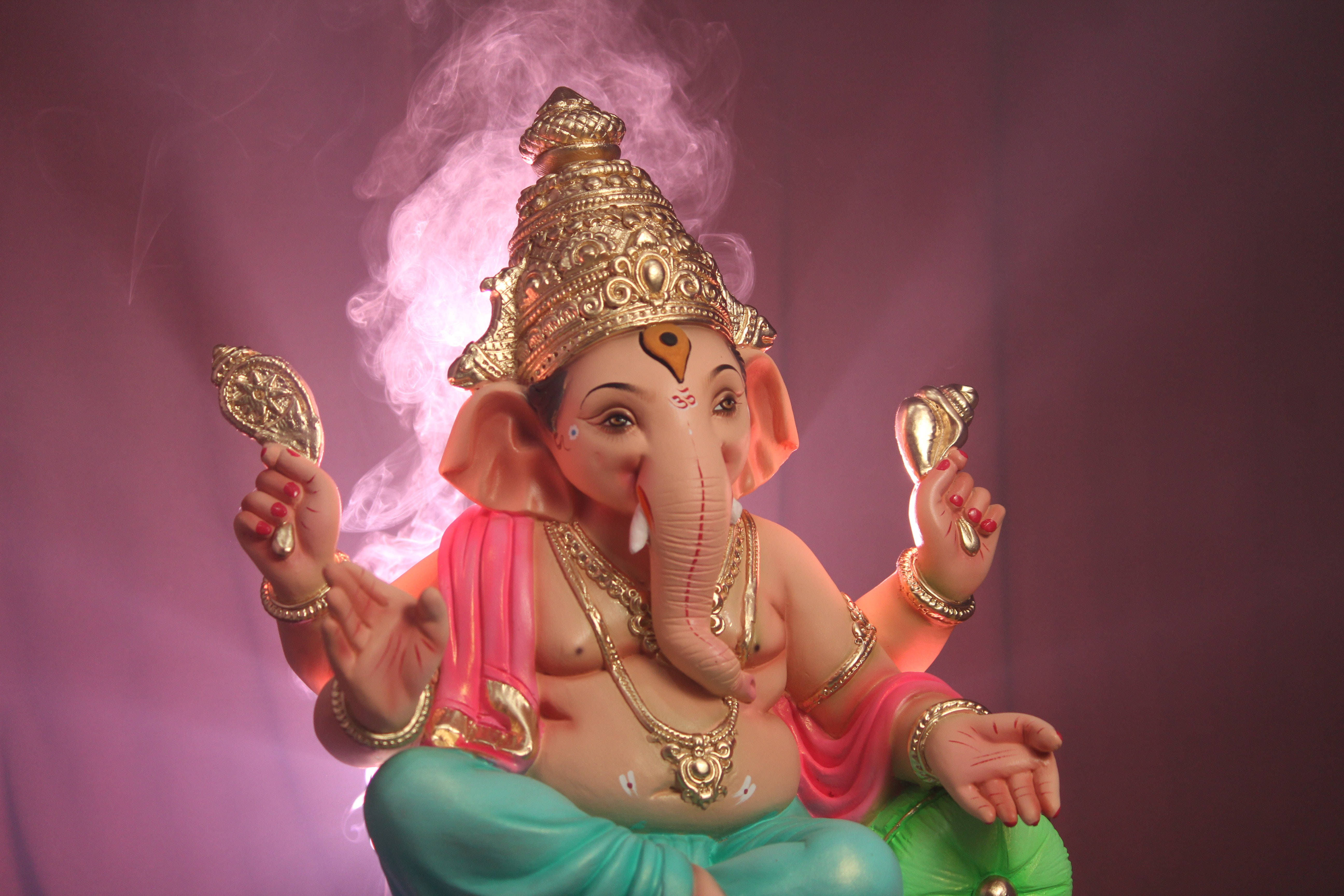 ganesh 4K 桌布 壁紙 — 一尊金色象神雕像，飾有珠寶和粉紅色布料，在煙霧瀰漫的紫色背景下安然端坐。