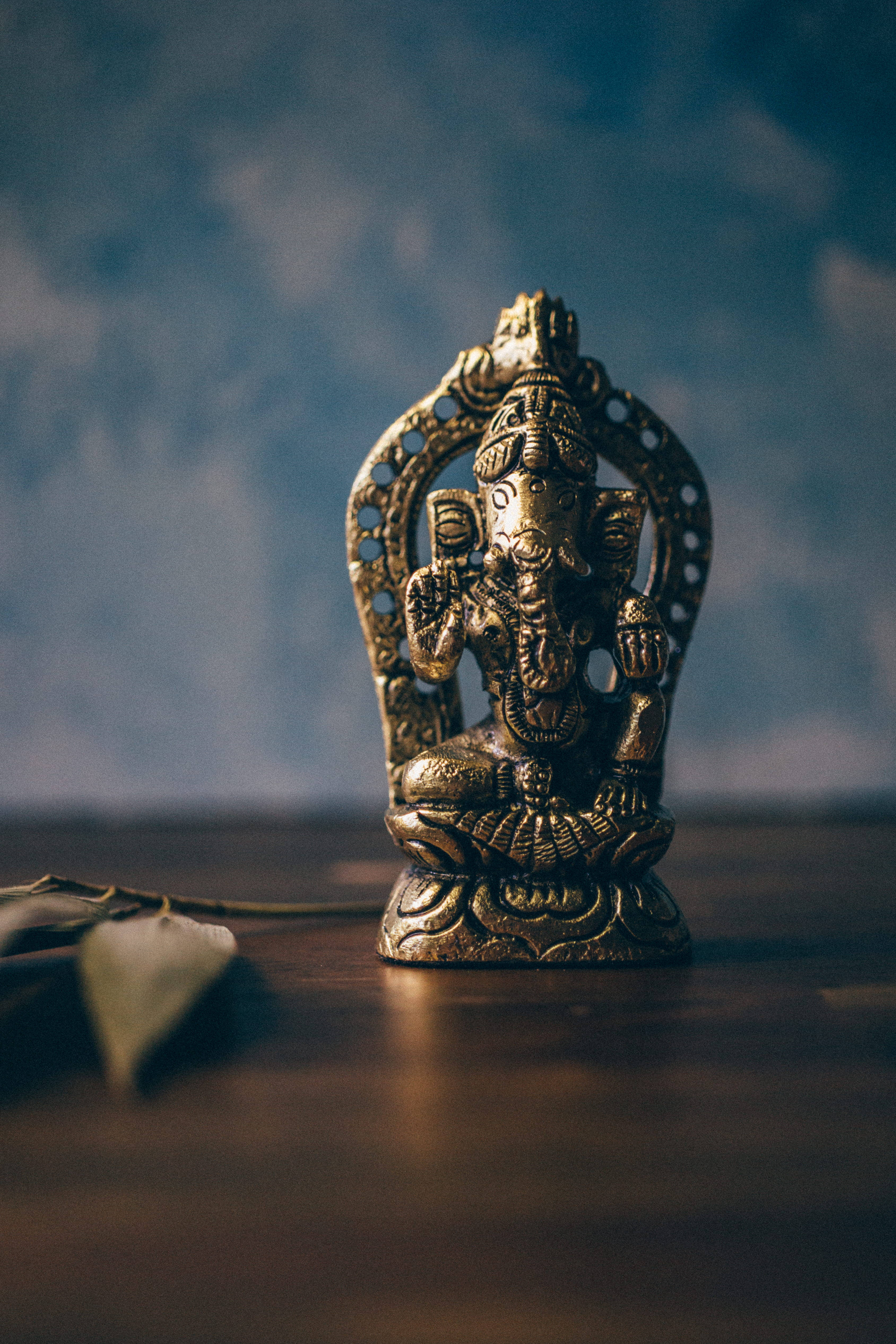 ganesh iPhone 桌布 壁紙 — 一尊金色象神雕像安詳地坐在木質表面上，背景是模糊的藍色，附近有一片葉子。