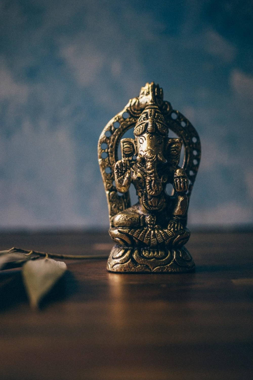 hindu iPhone 桌布 壁紙 — 一尊金色象神雕像安詳地坐在木質表面上，背景是柔和的藍色紋理。