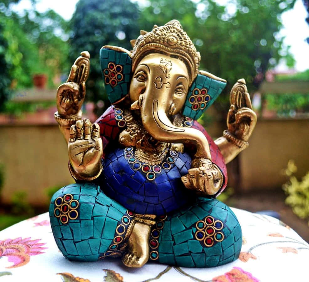Ganesh 桌布 壁紙 — 一尊金色馬賽克象神雕像，帶有綠松石和紅色點綴，安詳地坐在戶外的花卉坐墊上。