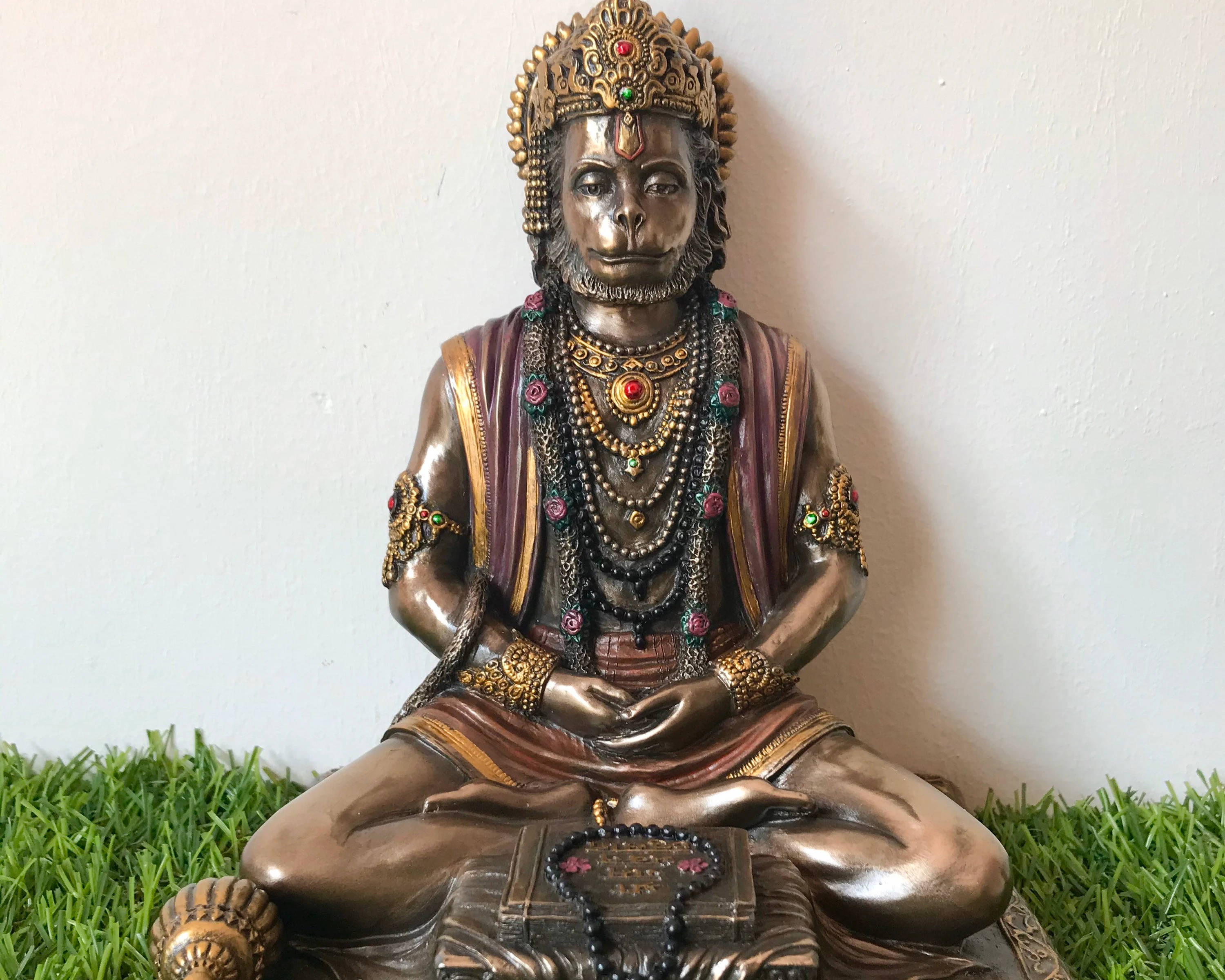 桌布 壁紙 — 一尊鑲有寶石的猴神青銅像，盤腿坐在寶座上，下方有綠草，背景是素牆。