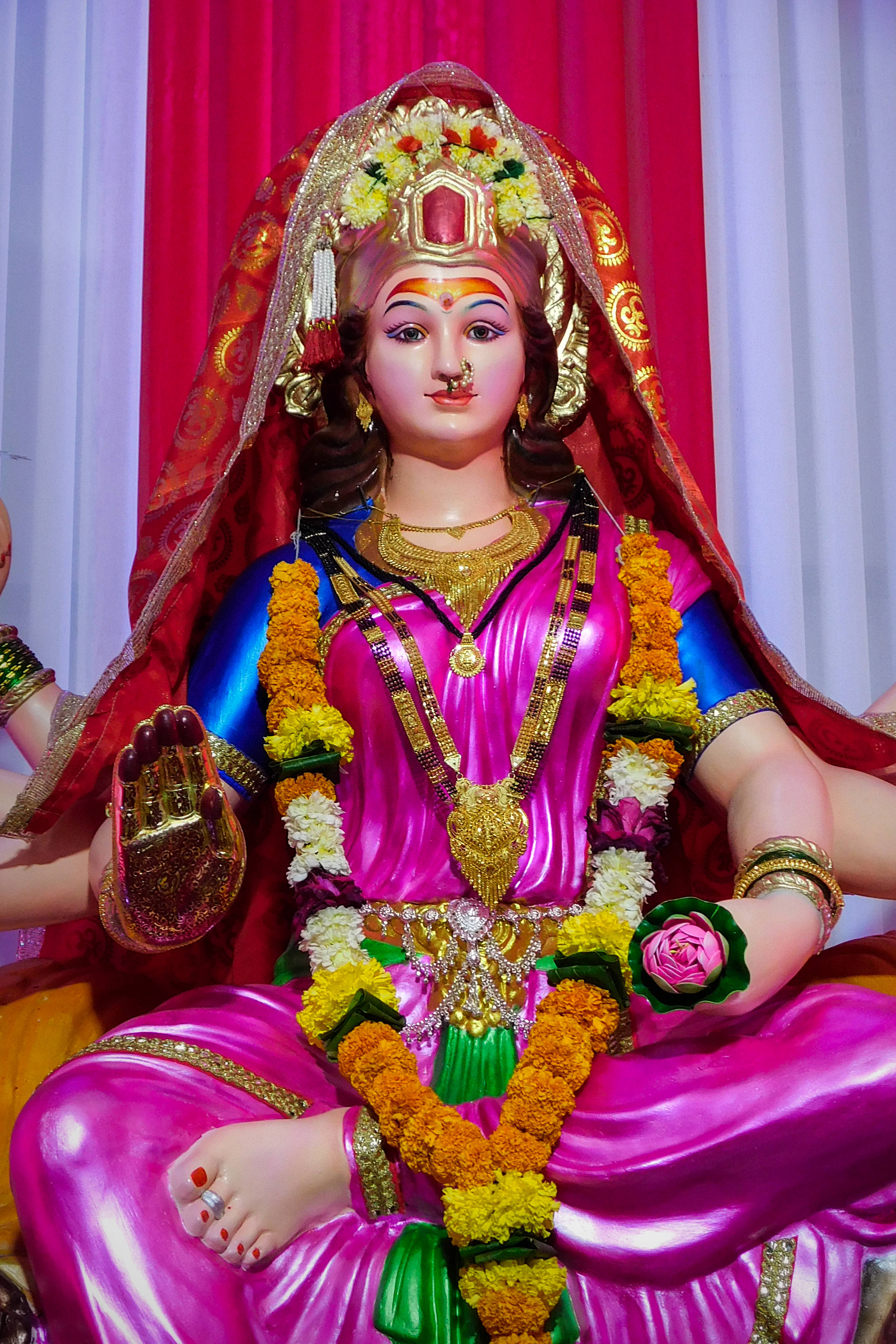maa durga 桌布 壁紙 — 一尊閃耀的印度教女神雕像，以粉紅和金色裝飾，佩戴萬壽菊花環，安詳地坐在紅色和白色帷幕前。