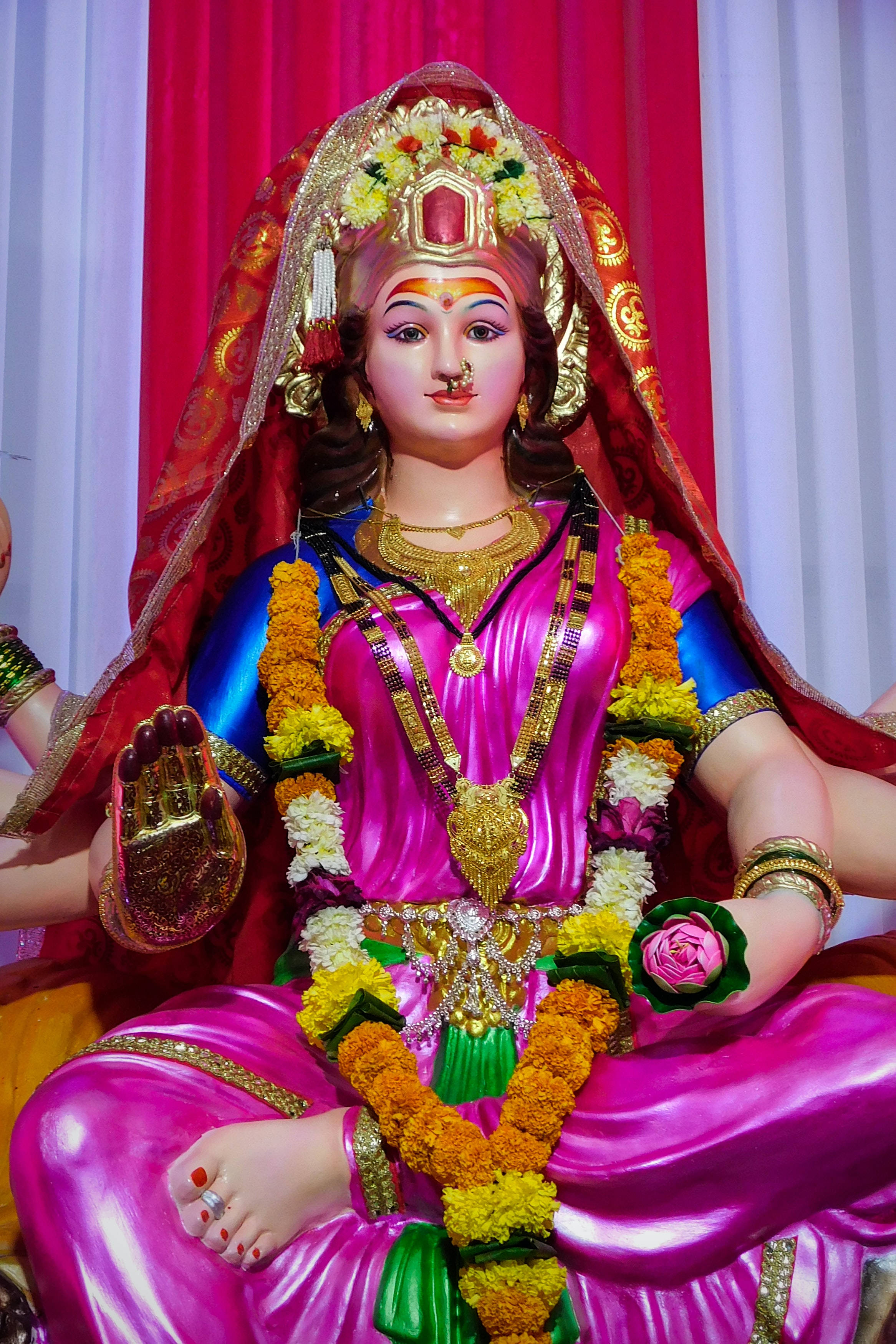 Mata Rani 桌布 壁紙 — 一尊閃耀的印度教女神雕像，以粉紅和金色裝飾，戴著萬壽菊花環，寧靜地坐在紅色和白色帷幕前。