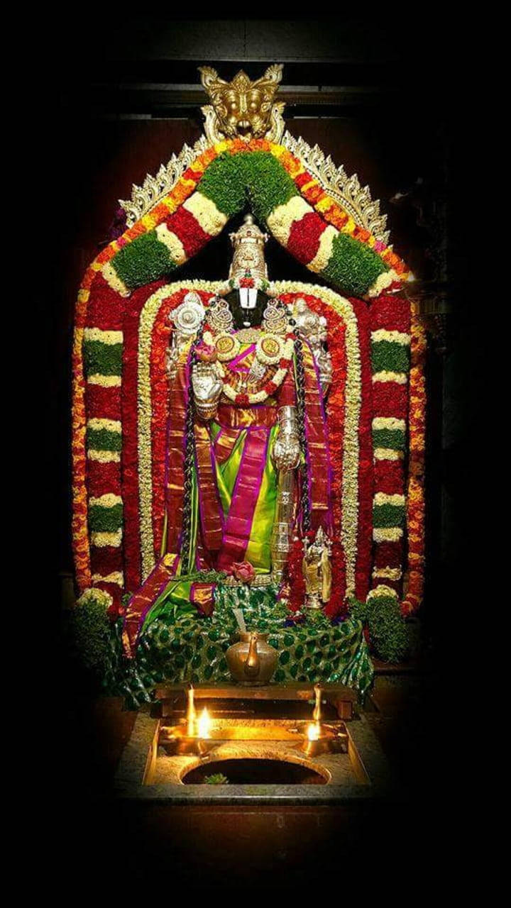 Lord Balaji 桌布 壁紙 — 一尊閃耀的印度教神像，飾以鮮豔的花環和金色飾物，在黑暗寺廟環境中被閃爍的油燈照亮。