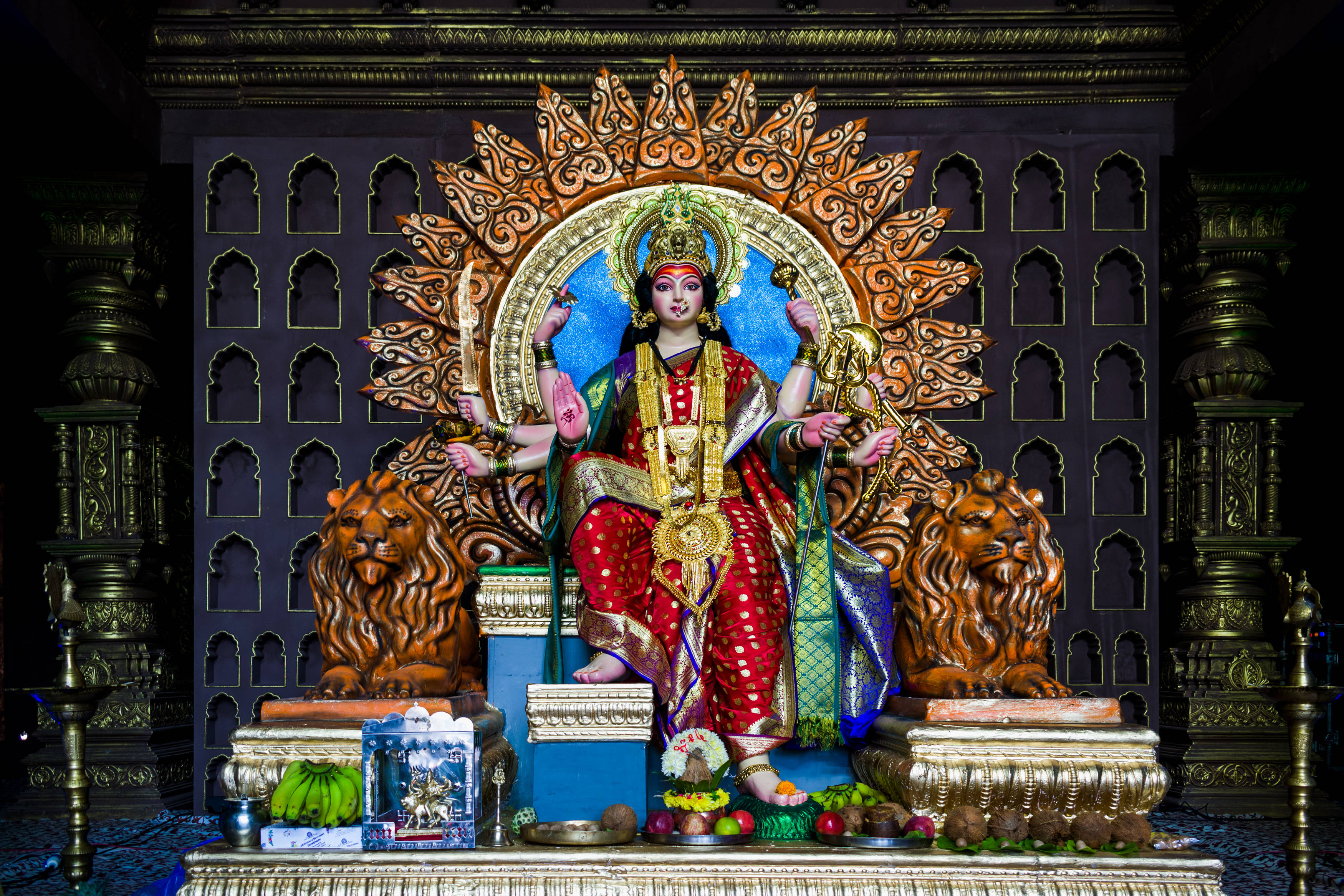 Durga 桌布 壁紙 — 一尊閃耀的多臂女神雕像，身穿紅金色，坐在兩頭獅子之間的寶座上，上方有華麗的太陽光環。
