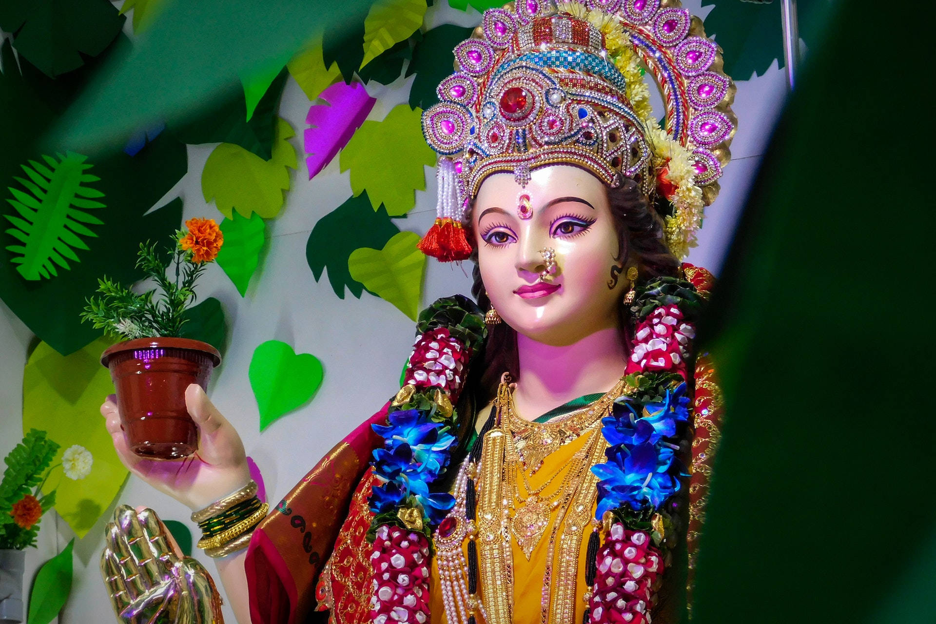 Durga 桌布 壁紙 — 一尊閃耀的神像，飾有珠寶和鮮花，手持一盆萬壽菊，背景是充滿活力的綠色葉子。