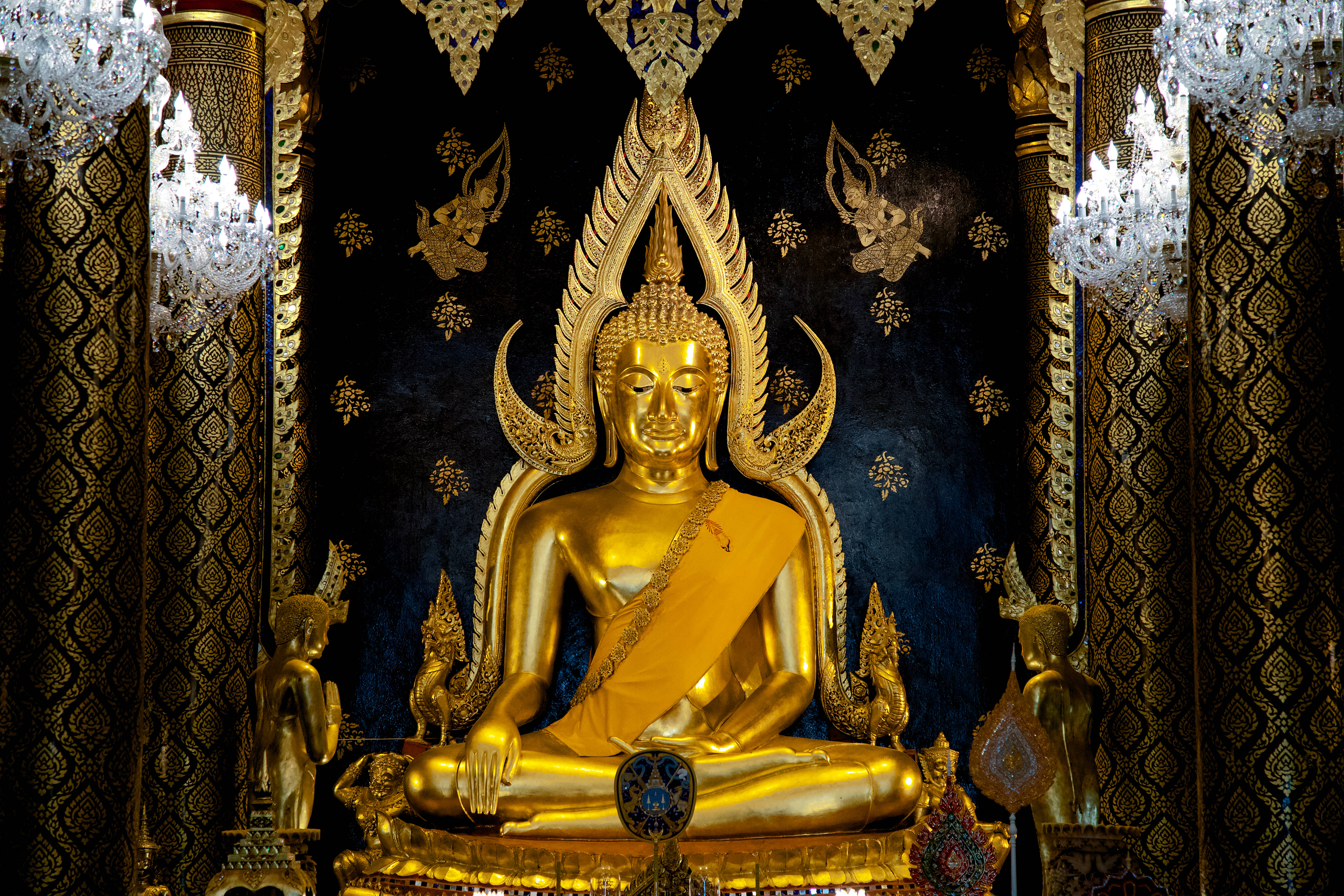 Gautam Buddha 桌布 壁紙 — 一尊閃耀的金色佛像靜靜地坐在裝飾華麗的寺廟神龕中，周圍有華麗的吊燈和深色圖案牆壁。