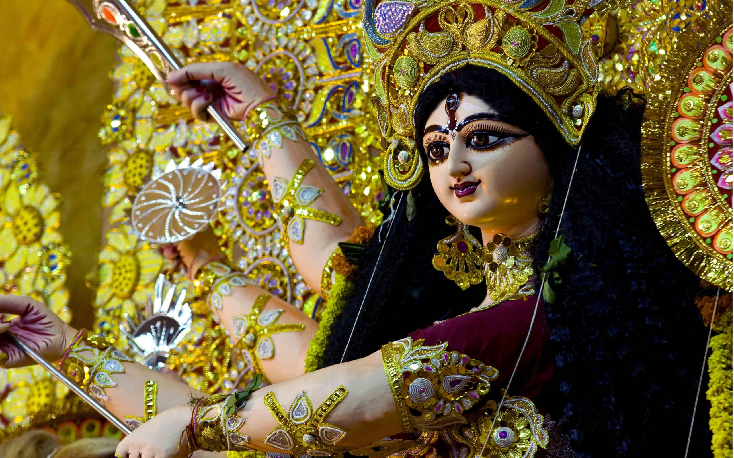 maa durga 桌布 壁紙 — 一尊閃耀的金色杜爾迦雕像，飾有精緻珠寶，手持武器，背景是鮮豔的花卉。