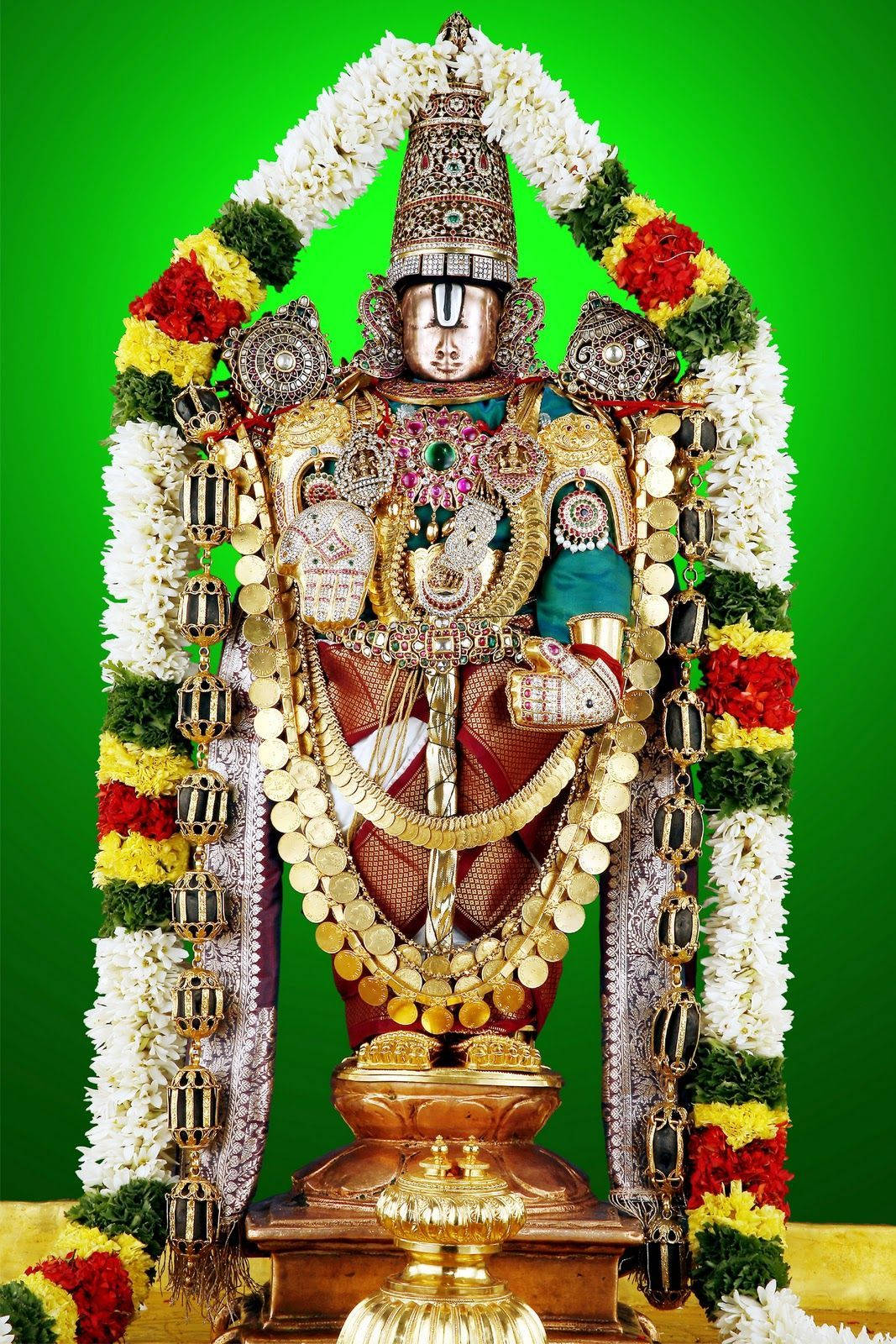 Venkateswara 桌布 壁紙 — 一尊閃耀的金色神像，飾以精緻珠寶和色彩繽紛的花環，襯托著充滿活力的綠色背景。