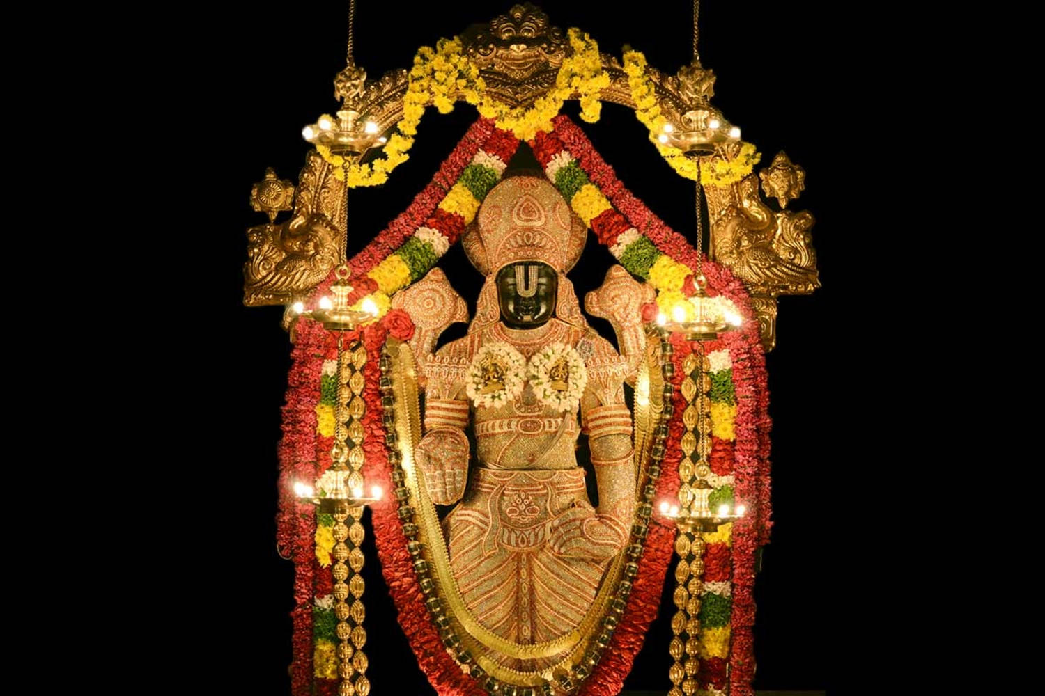 Vishnu 桌布 壁紙 — 一尊閃耀的金色神像，飾以鮮豔的紅、黃、綠色花環和發光的油燈，背景為黑色。