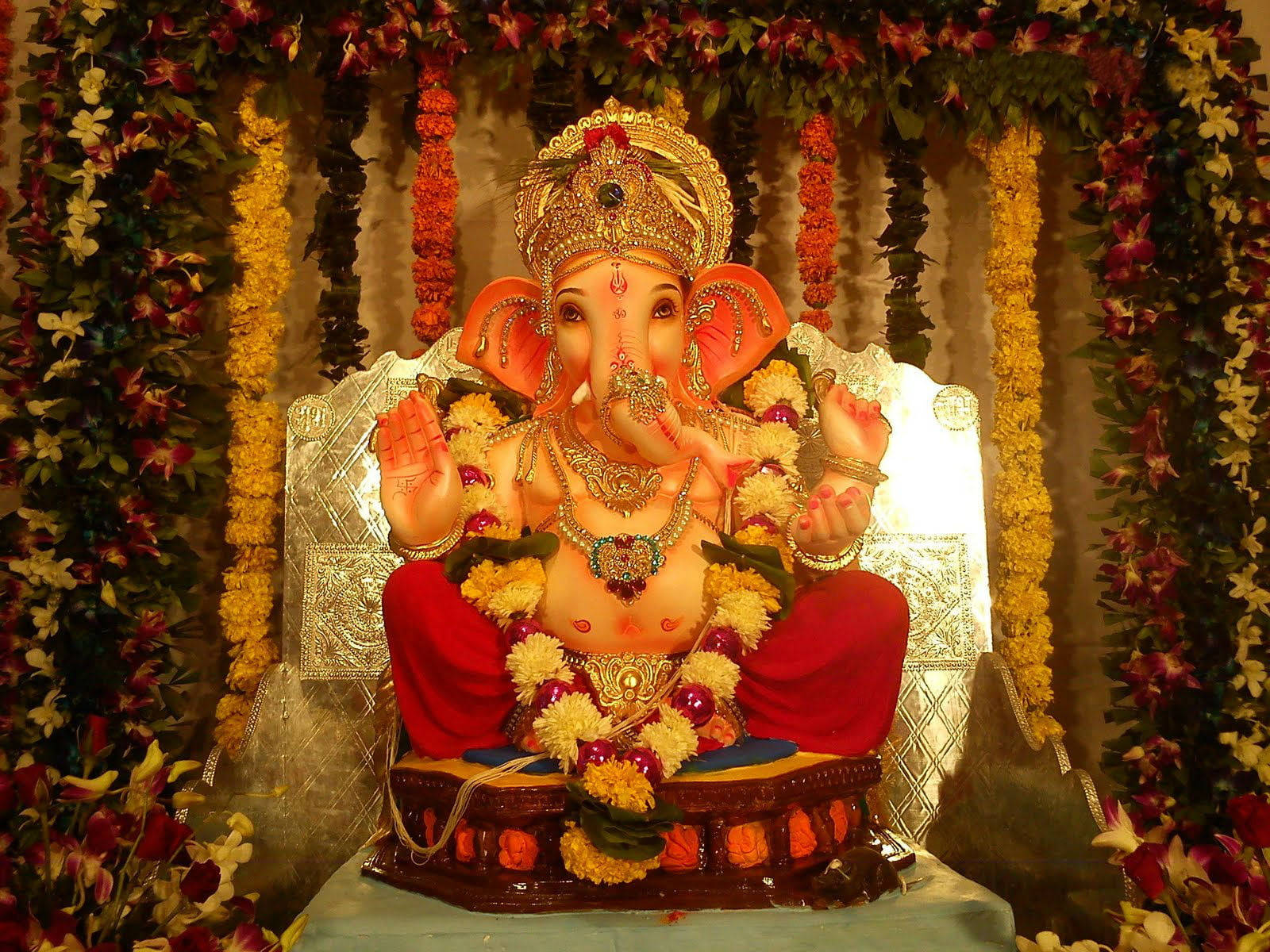 Ganesh 桌布 壁紙 — 一尊閃耀的金色象神像，飾以紅黃花朵，安詳地坐在裝飾華麗的基座上。