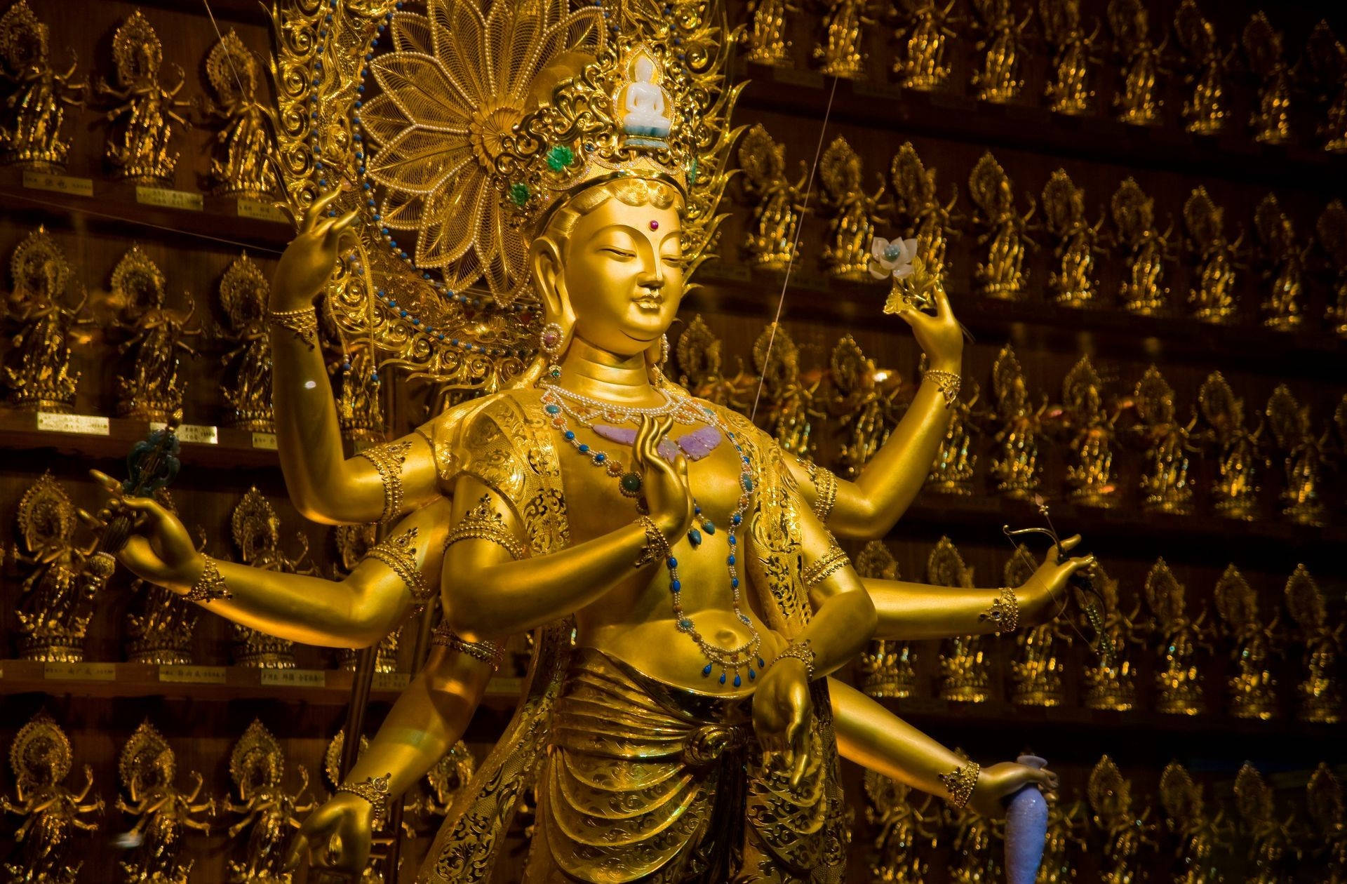 maa durga 桌布 壁紙 — 一尊閃耀金色多臂佛像，飾有珠寶，立於寺廟內擺放較小金佛的架子前。