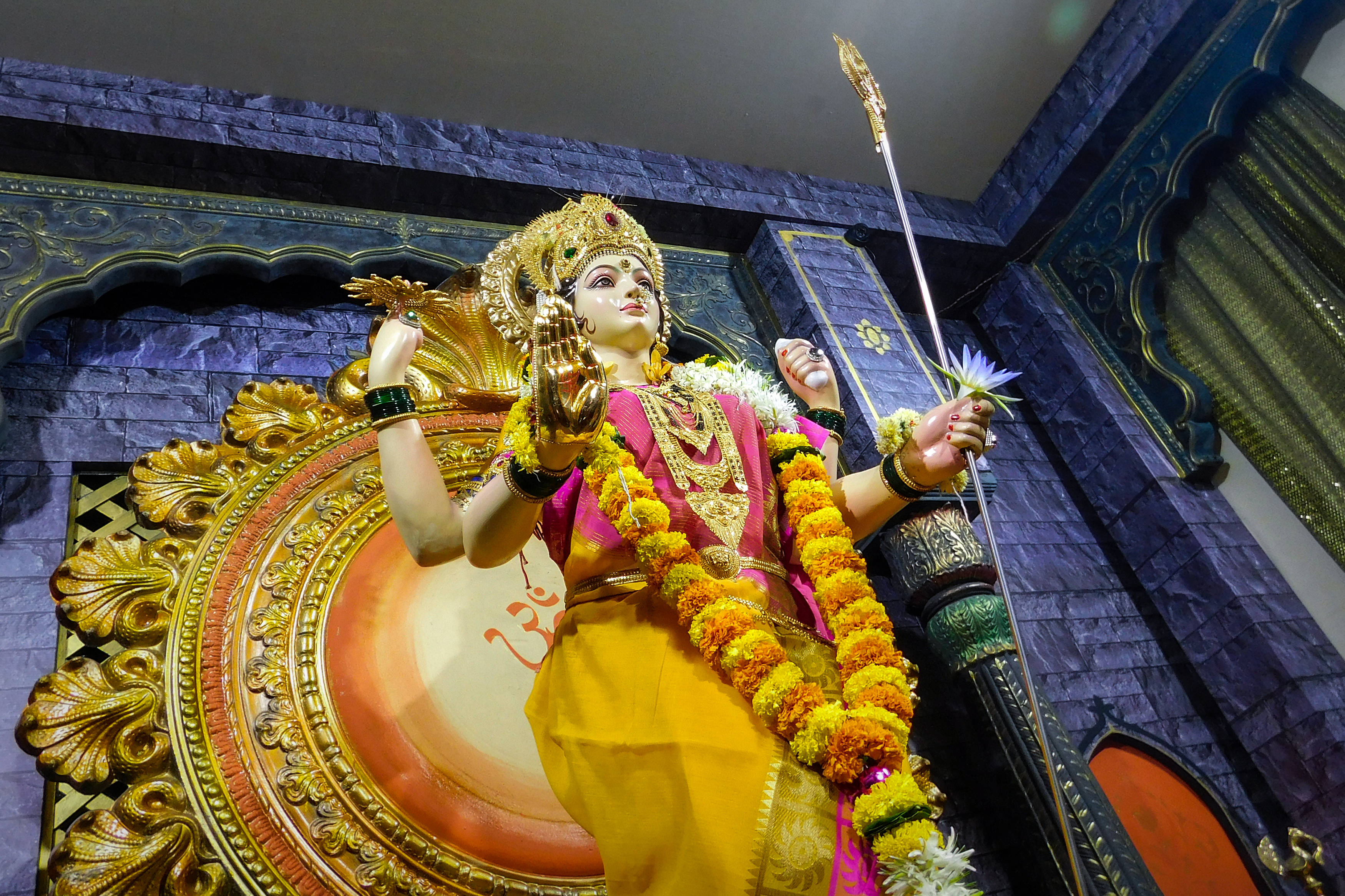 Navratri 桌布 壁紙 — 一尊閃閃發光的金色印度教女神雕像，裝飾著萬壽菊花環，手持劍，站在華麗的藍金色寺廟背景前。