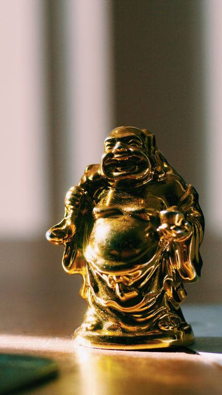 Statue Aesthetic 桌布 壁紙 — 一尊閃閃發光的金色笑佛雕像，坐在沐浴在溫暖陽光下的木質表面上，背景柔和模糊。
