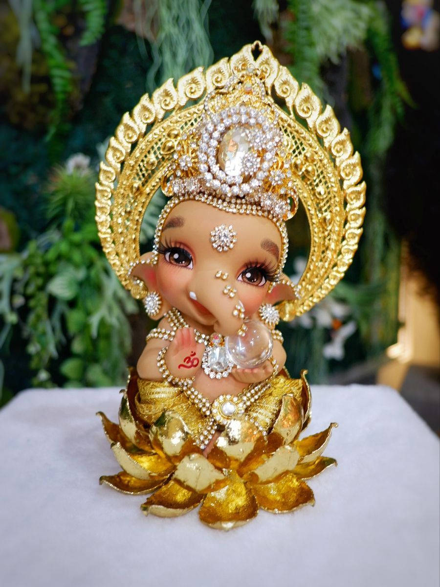 cute ganesha 桌布 壁紙 — 一尊閃閃發光的金色象神雕像，飾有珠寶，優雅地坐在蓮花底座上，背景是茂密的綠色樹葉。