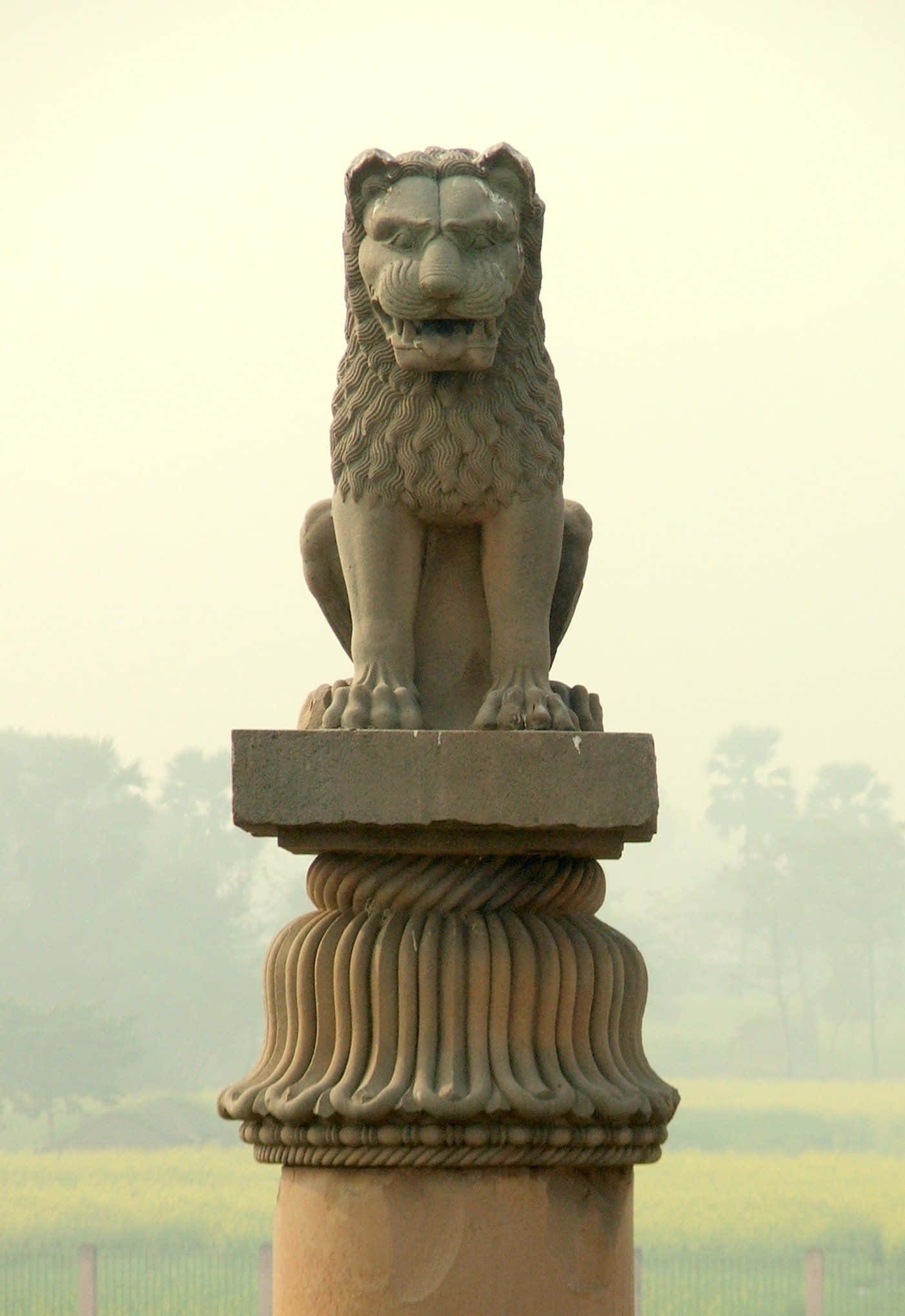 statue 桌布 壁紙 — 一尊雄偉的石獅雕像棲息在華麗的柱子上，背景是朦朧的樹木和黃色田野。