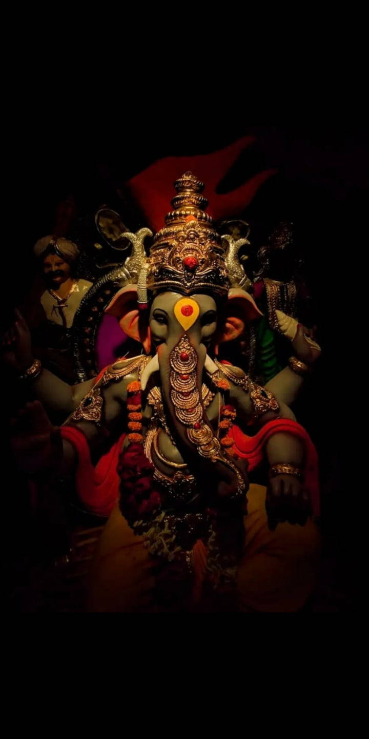 hindu iPhone 桌布 壁紙 — 一尊雄偉的金色裝飾象神雕像，在黑暗背景中照亮，配有紅色布料和背後微笑的侍者。
