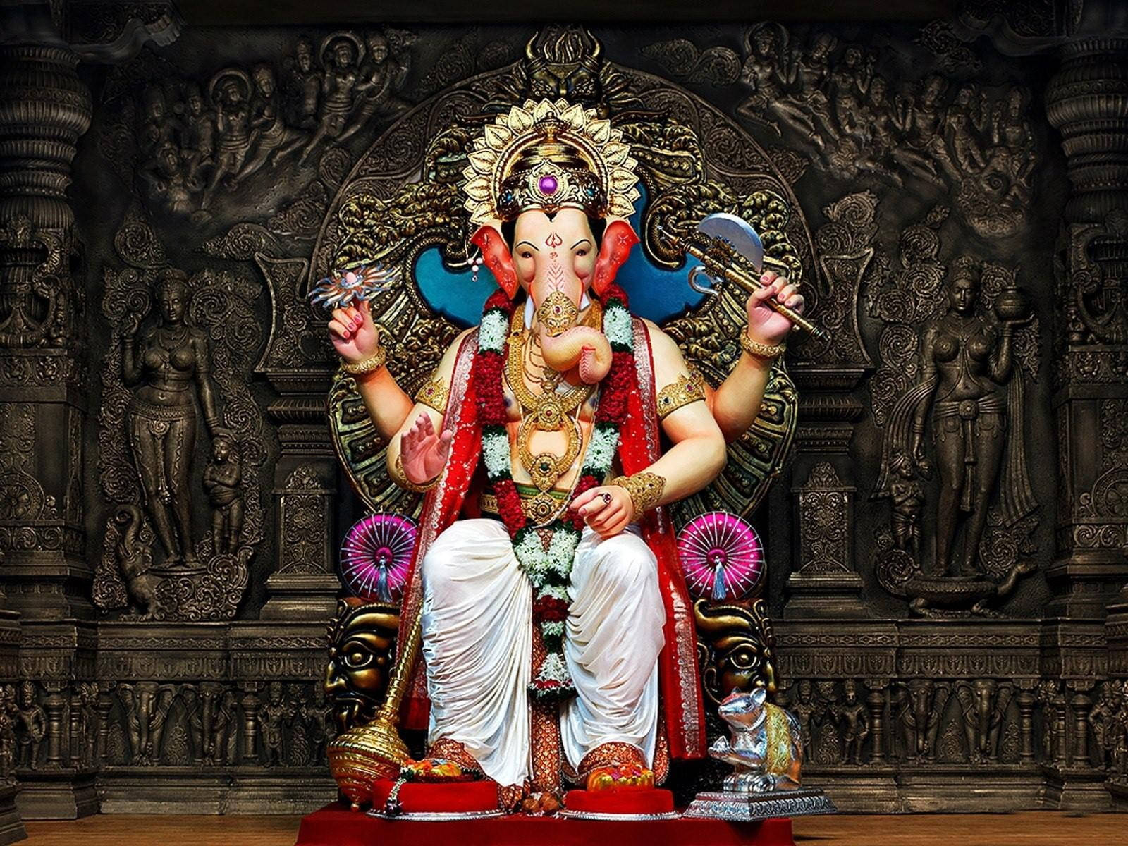 Ganesh 桌布 壁紙 — 一尊雄偉的金色象神像，飾以紅色花環和珠寶，威嚴地坐在華麗的寶座上，背景是深色雕刻的寺廟。