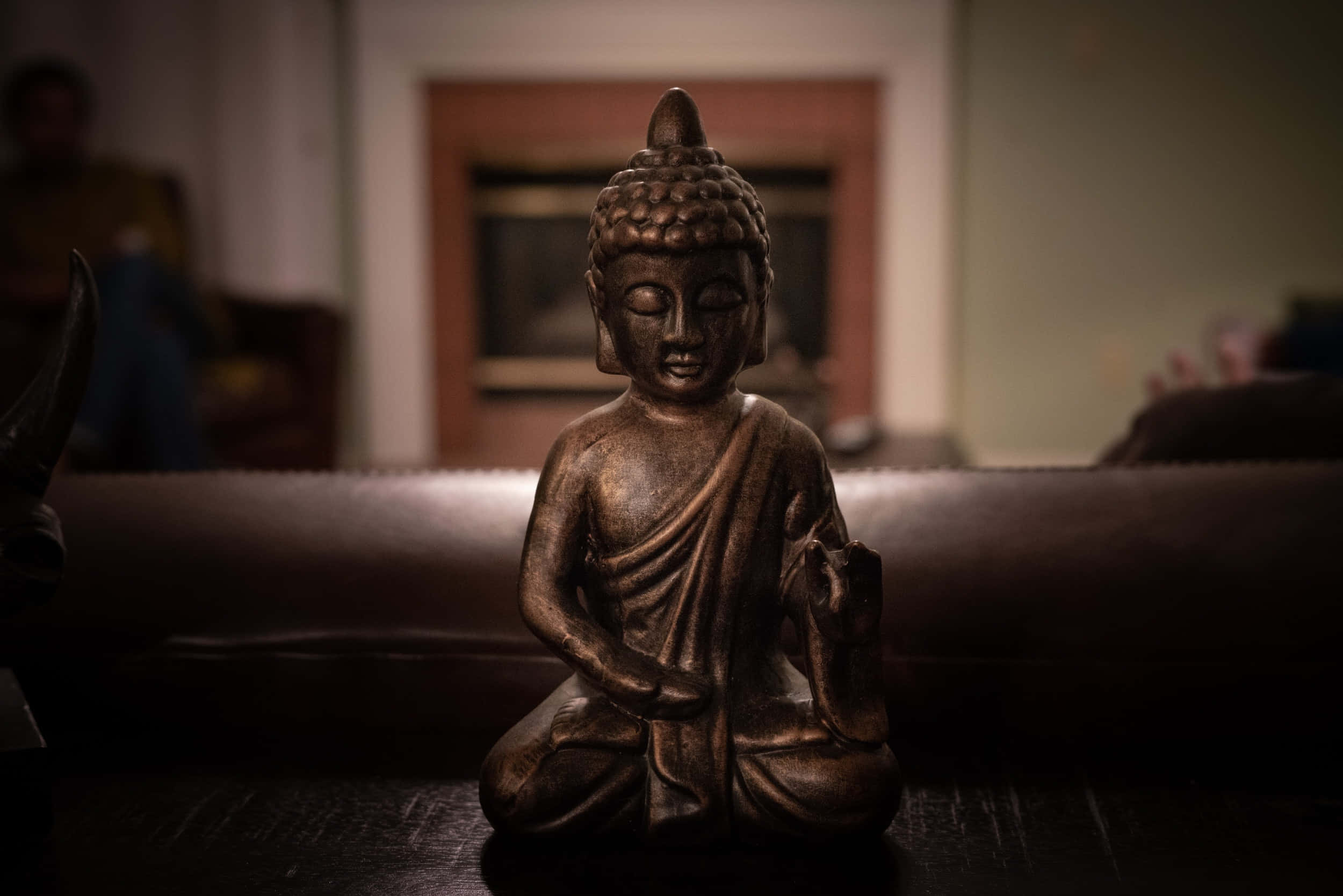 佛陀 桌布 壁紙 — 一尊青銅佛像在光線昏暗的房間中靜坐，背景家具柔和模糊。