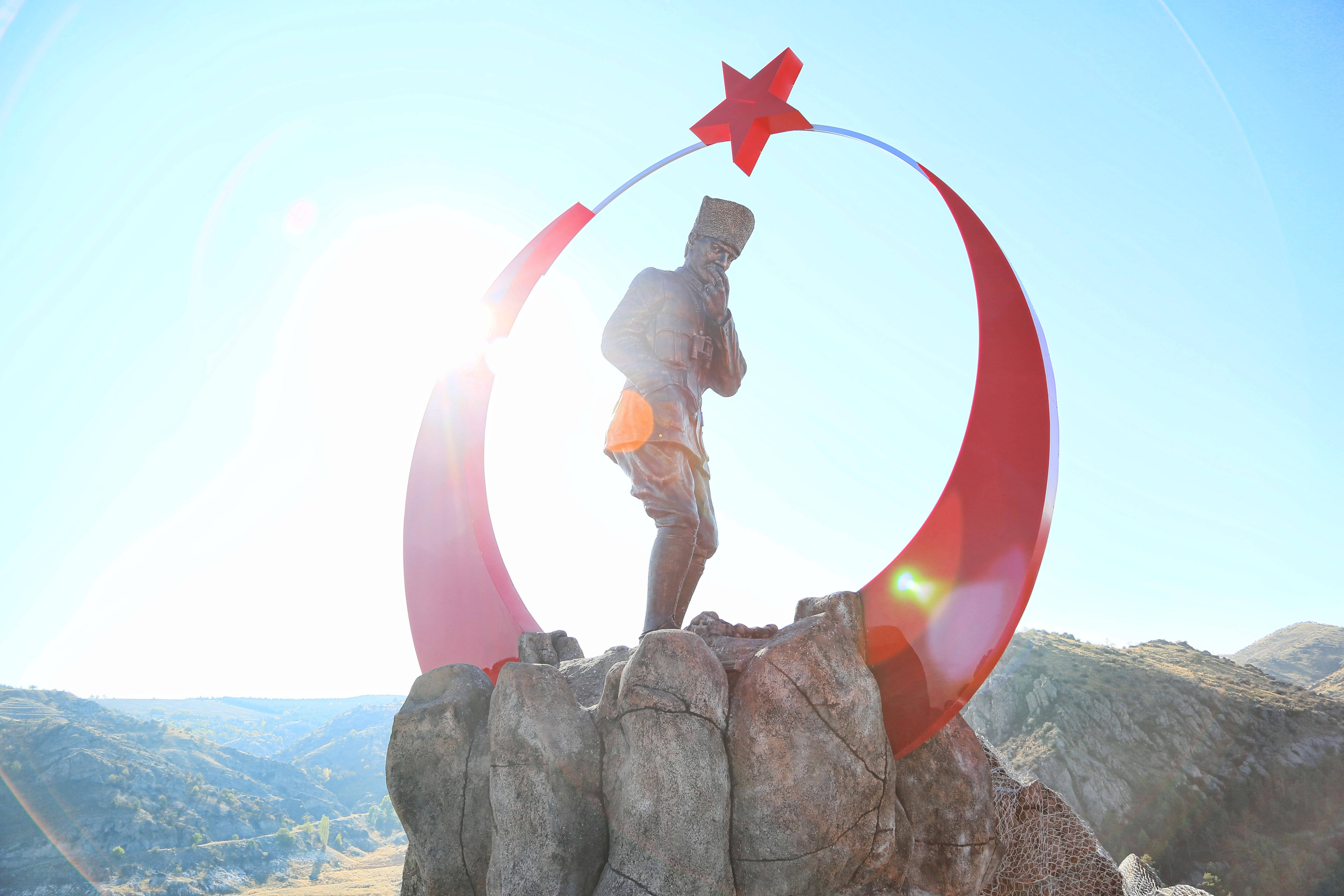 Ataturk 桌布 壁紙 — 一尊青銅士兵雕像矗立在岩石地形上，以紅色星星和新月拱門為框，映襯著明亮的陽光天空。