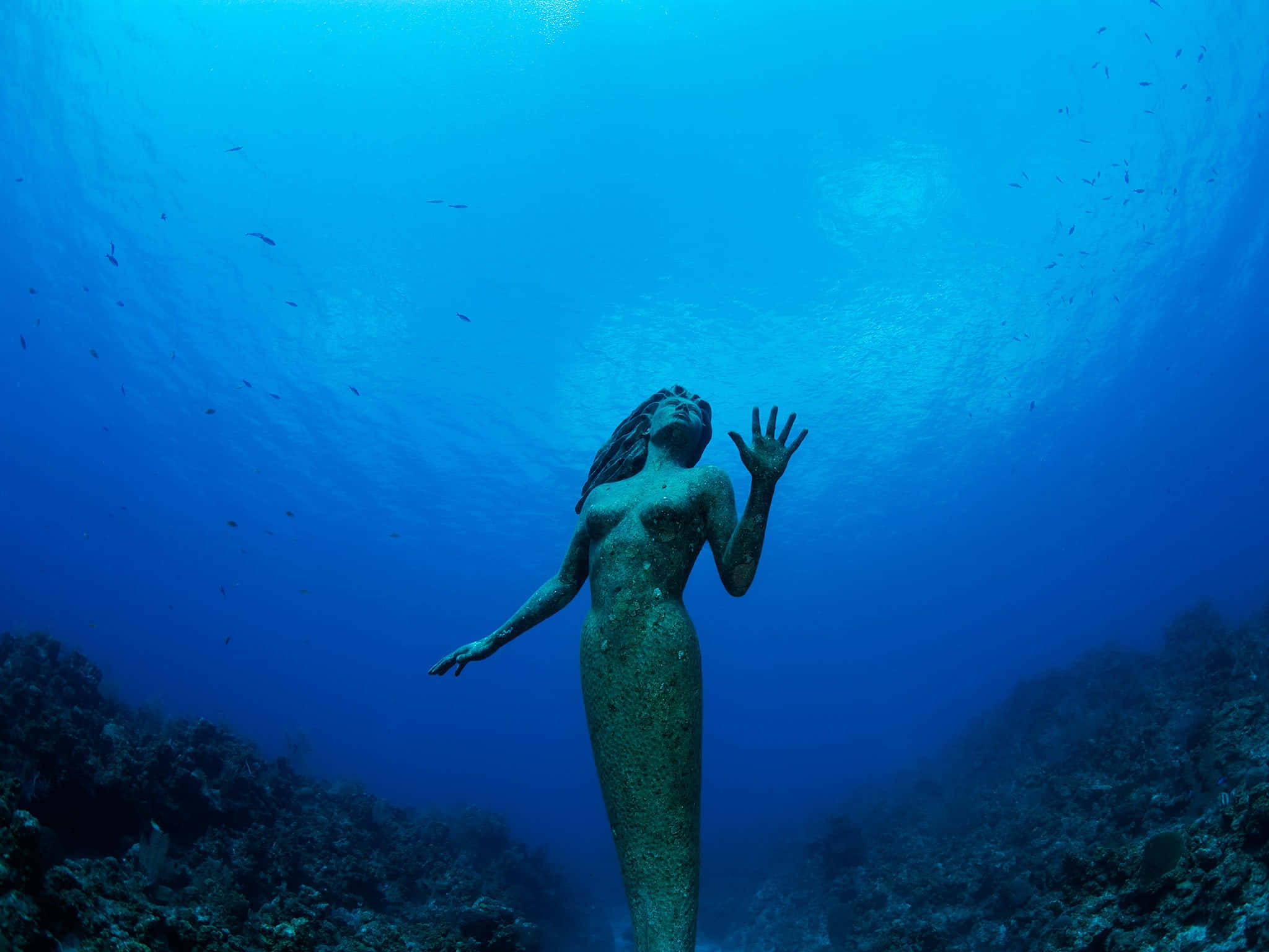 Underwater Ocean 桌布 壁紙 — 一尊青銅美人魚雕像在水中優雅地張開雙臂站立，陽光穿過上方深藍色的海洋照射下來。