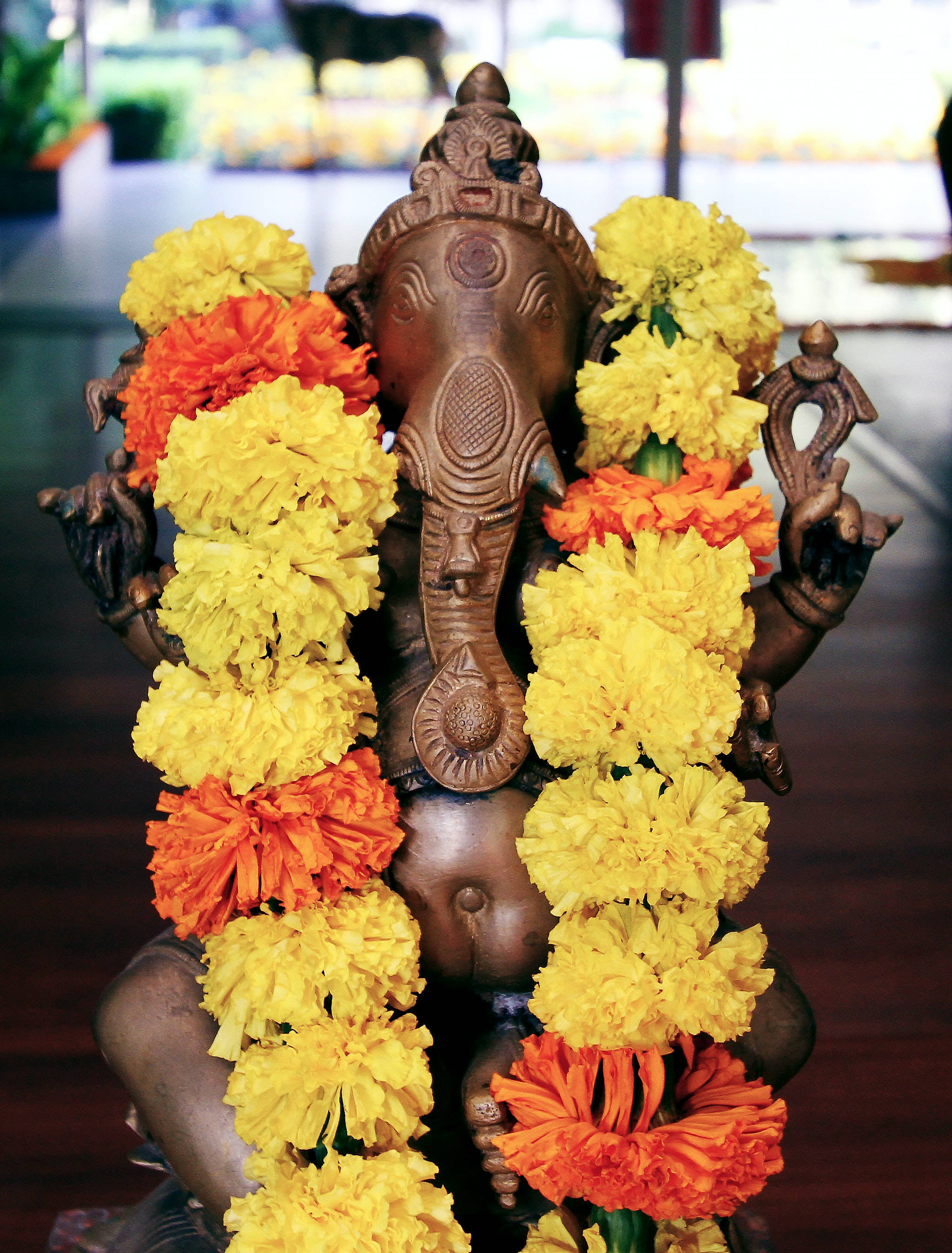 Ganesh 桌布 壁紙 — 一尊青銅象神雕像，飾以鮮豔的黃色和橙色萬壽菊花環，置於室內，背景模糊。