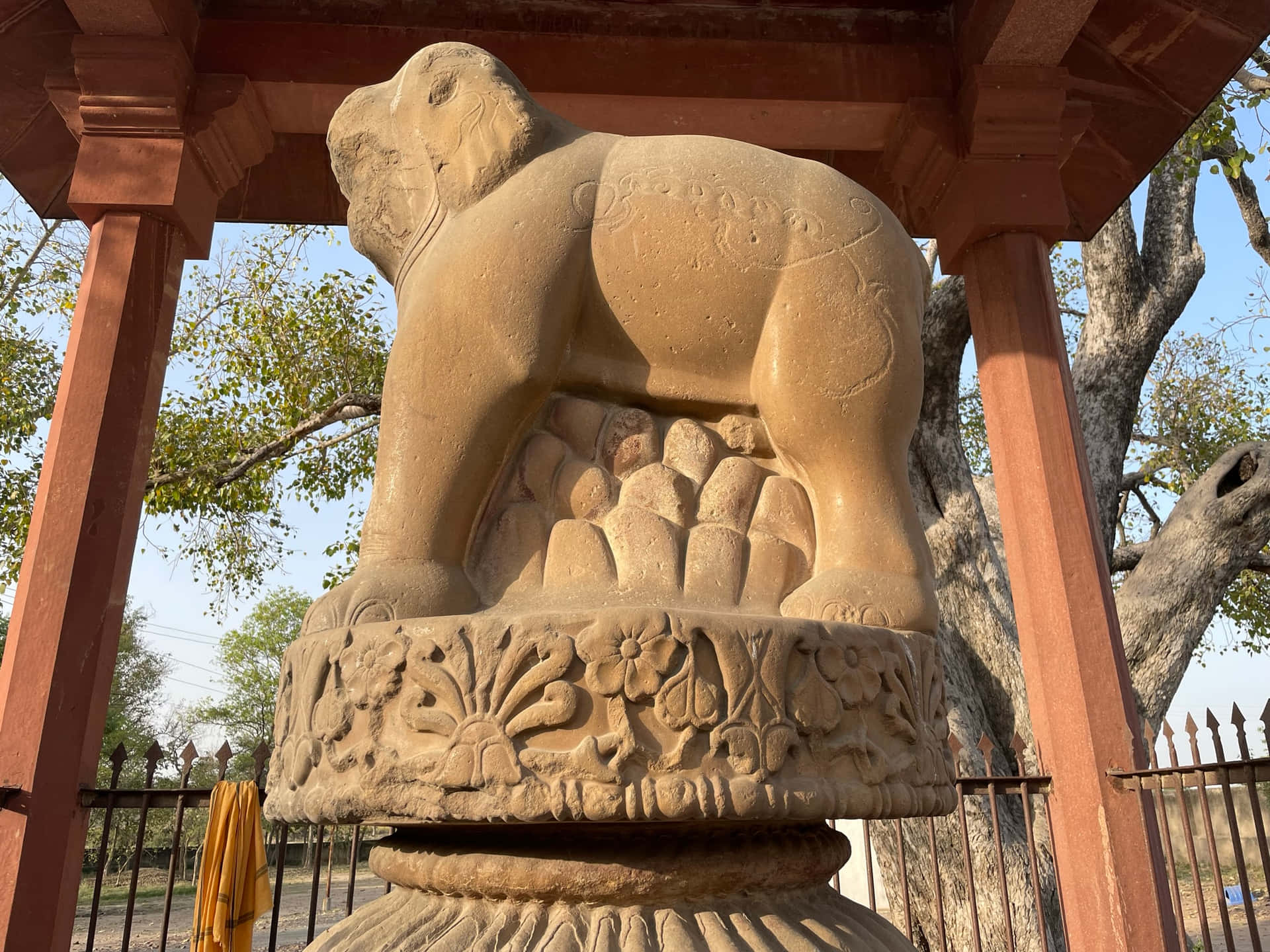 ASHOKA PILLAR ELEPHANT 壁紙 — 一尊風化的石象雕像矗立在裝飾華麗的基座上，位於紅色木亭下，背景有樹木和天空。