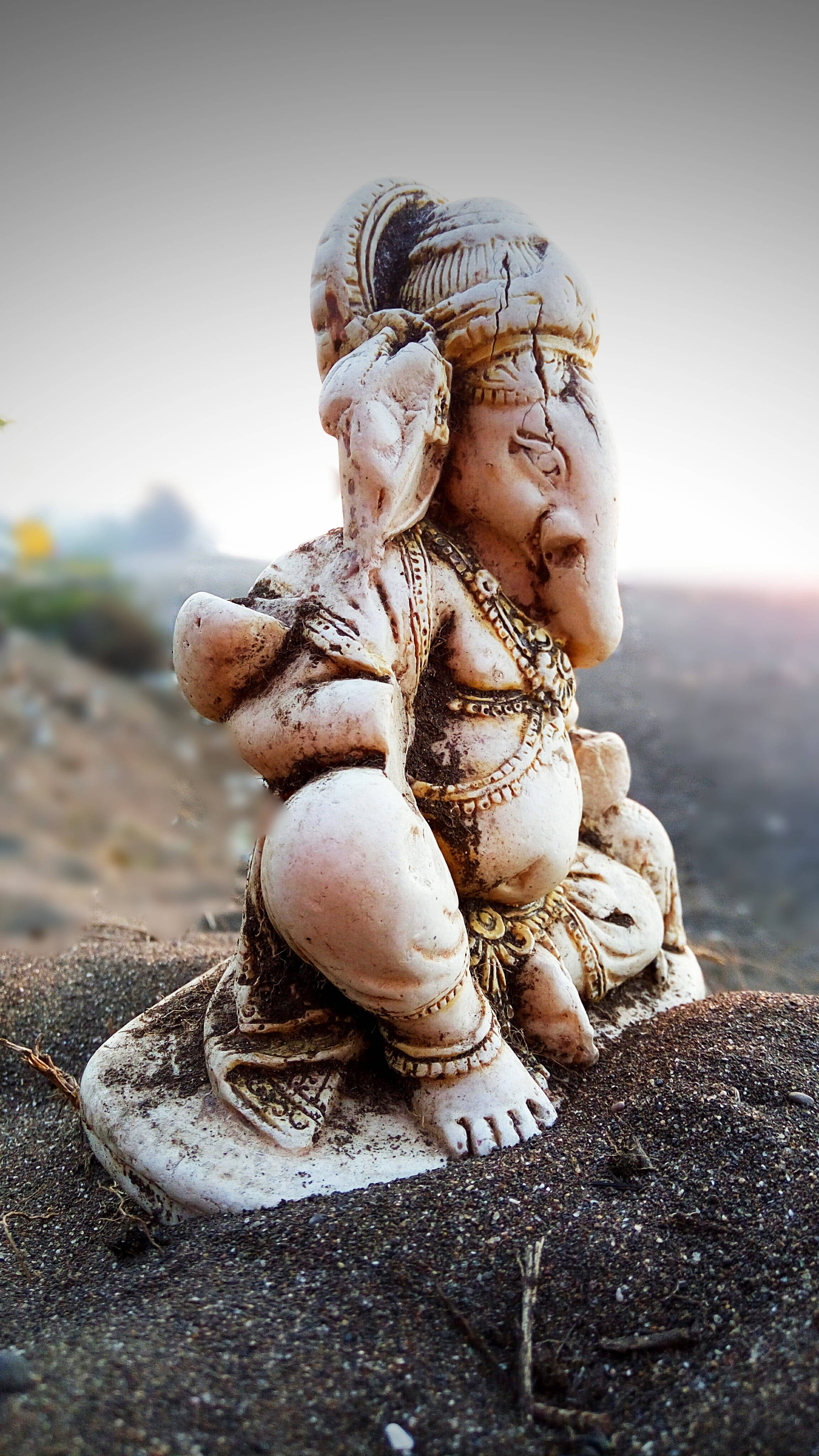 ganesh iPhone 桌布 壁紙 — 一尊風化的石雕象神雕像安詳地坐在深色泥土上，背後有柔和的金色光芒照耀。