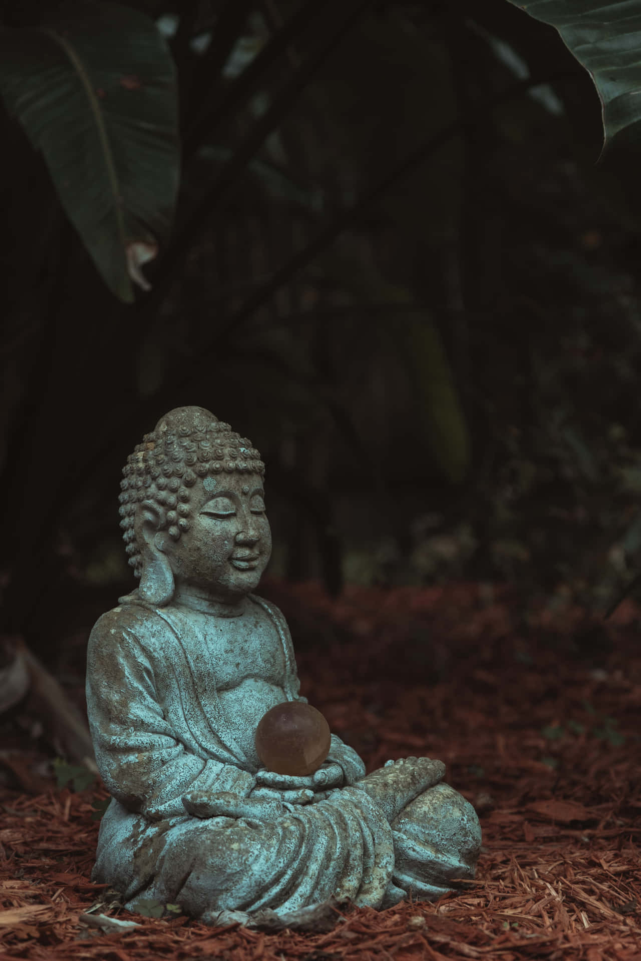 佛陀 桌布 壁紙 — 一尊風化的綠松石佛像，在大片綠葉下的豐富棕色覆蓋物中，平靜地坐著，手持水晶球。