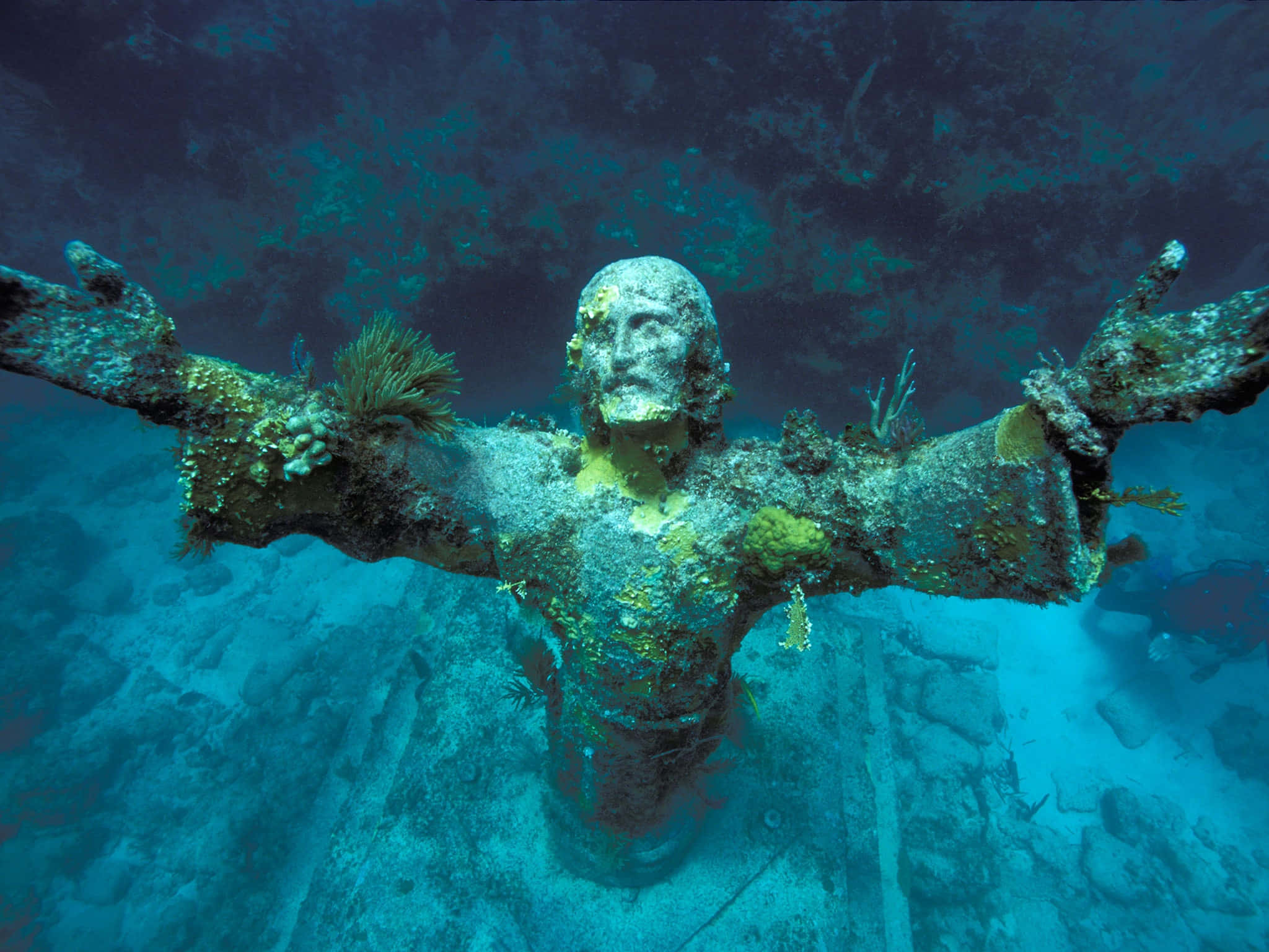 Underwater Ocean 桌布 壁紙 — 一尊風化的耶穌張開雙臂雕像浸沒在水中，在藍綠色水下覆蓋著珊瑚和海洋生物。