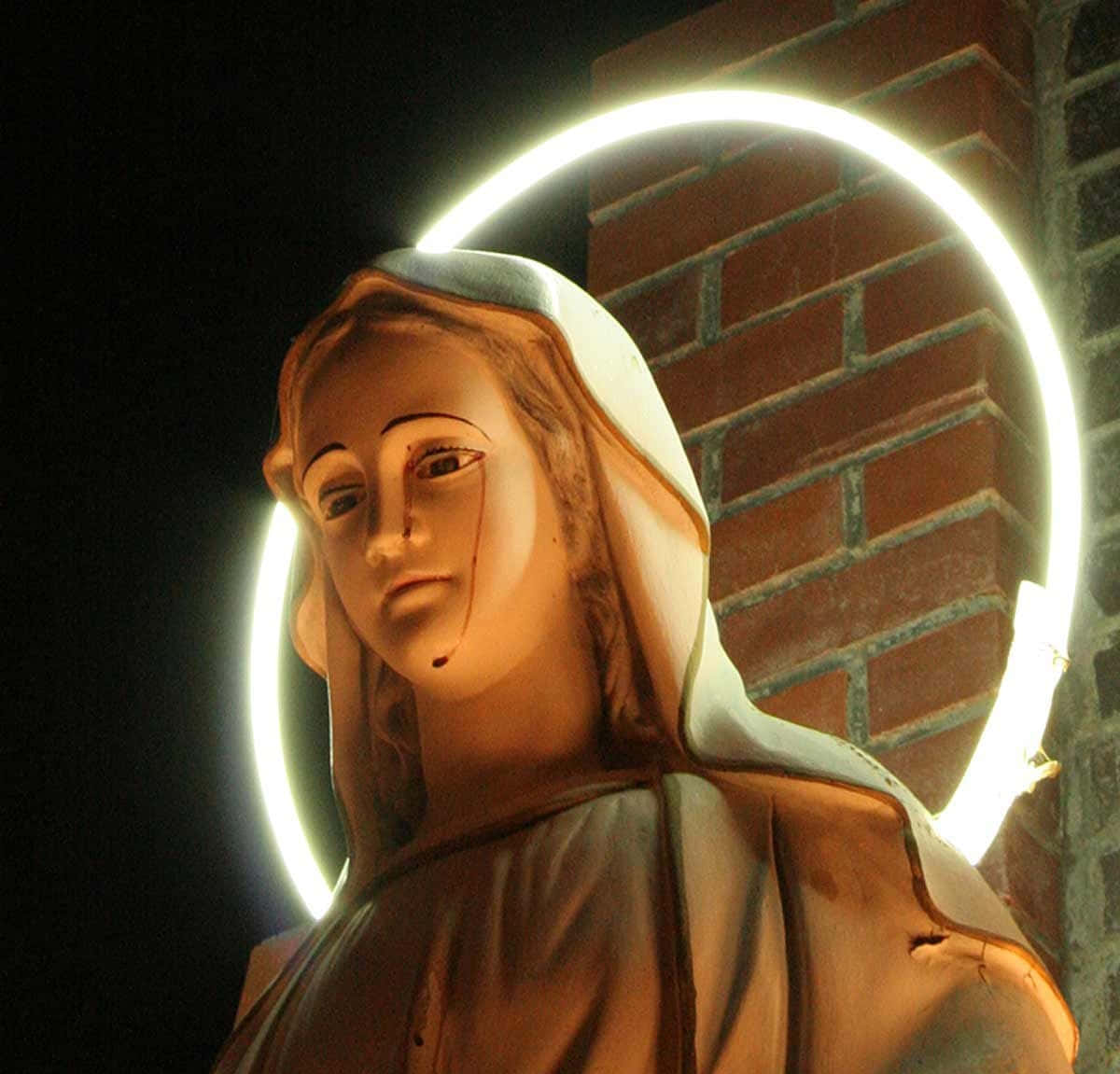 Mother Mary 桌布 壁紙 — 一尊風化的聖人雕像，帶有發光的光環，夜晚矗立在磚牆前。