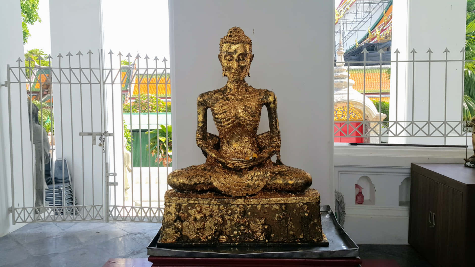 佛陀 桌布 壁紙 — 一尊風化的金色佛像安然坐在室內，以白色柱子和金屬門框為背景，外面可見茂密的綠植。