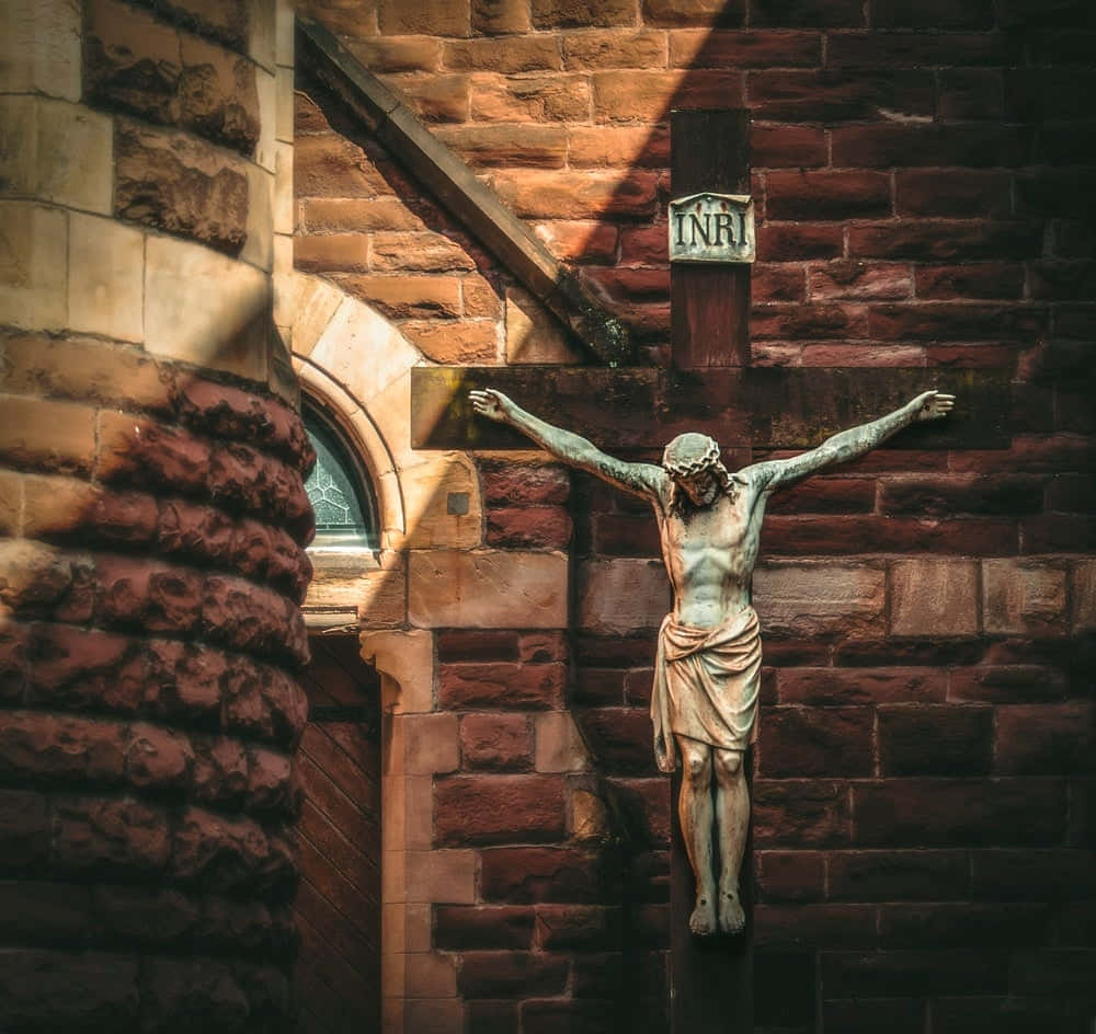 Jesus Crucifixion 桌布 壁紙 — 一尊風化的青銅十字架受難像，在溫暖陽光下，於質感豐富的紅磚教堂牆上投下長長的陰影。