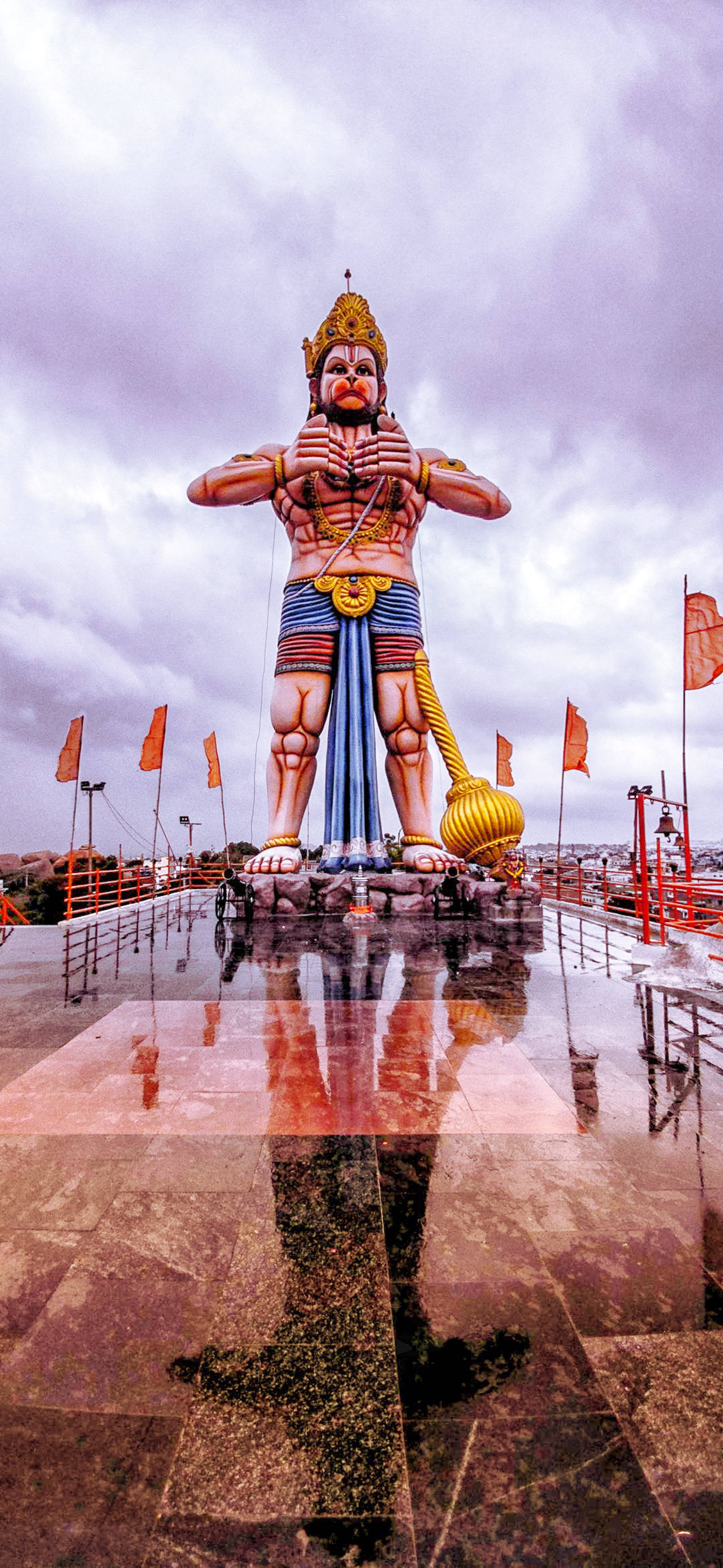 hanuman art 桌布 壁紙 — 一尊高聳的哈努曼神像威嚴地矗立在潮濕的平台上，四周是戲劇性的多雲天空和鮮豔的橙色旗幟飄揚。