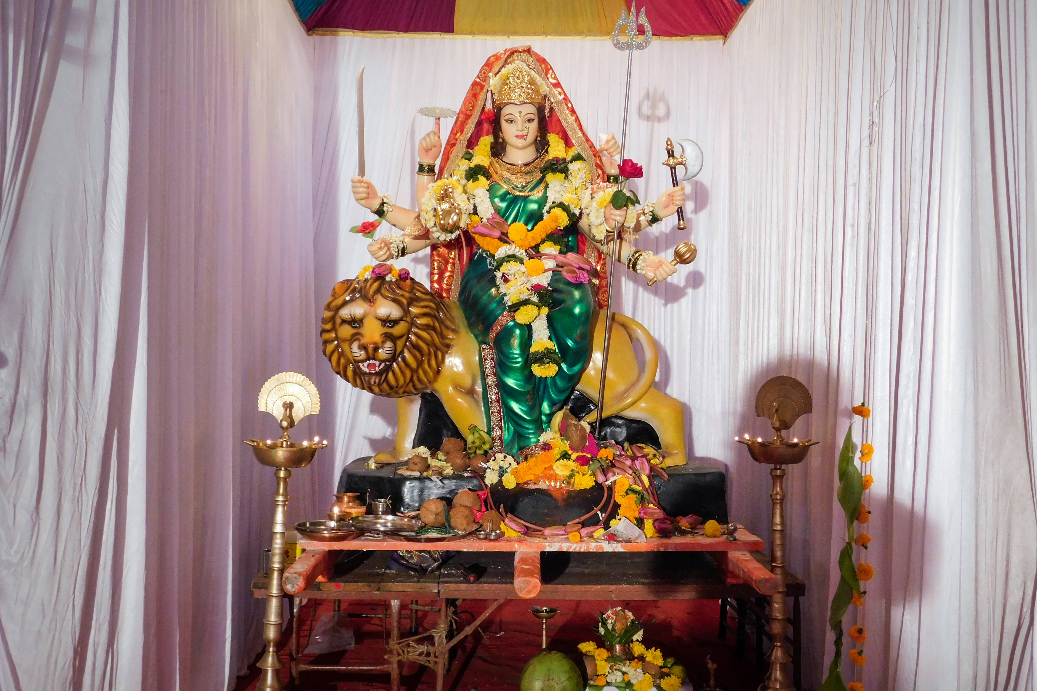 Navratri 桌布 壁紙 — 一尊鮮綠色、多臂印度教女神坐在獅子上的雕像，飾有鮮花，周圍環繞著儀式用品，下方有垂墜布料。
