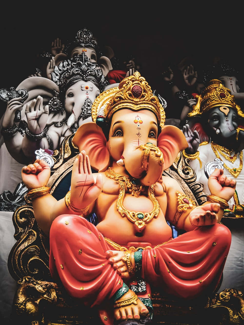 Ganesh 桌布 壁紙 — 一尊鮮豔的金色和紅色象頭神像顯眼地坐著，周圍環繞著其他穿著華麗服飾的象頭神像，背景為深色。