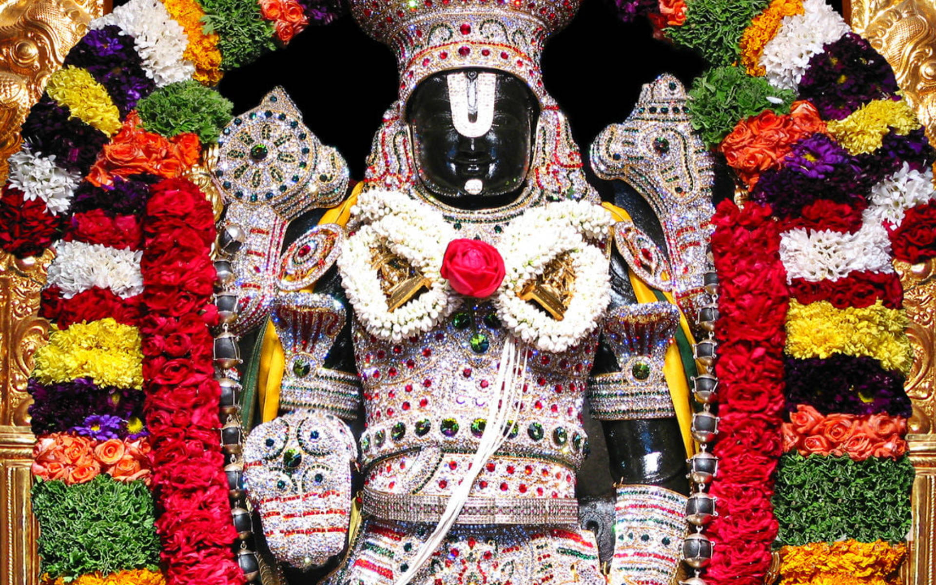 Lord Balaji 桌布 壁紙 — 一尊黑色神像，飾以閃爍的珠寶和紅、黃、綠色的鮮豔花環。