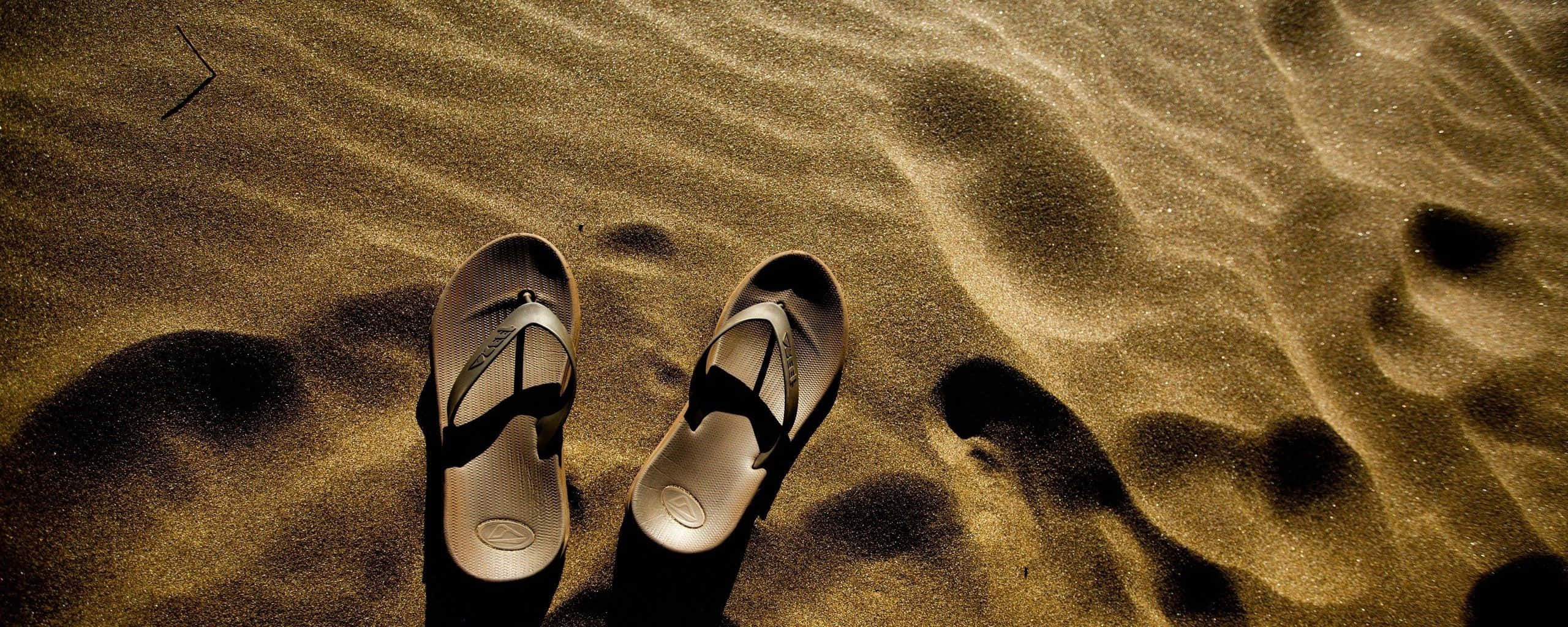 Beach Sandals 桌布 壁紙 — 一對人字拖放在波紋狀的金色沙灘上，溫暖的陽光投下深深的陰影。