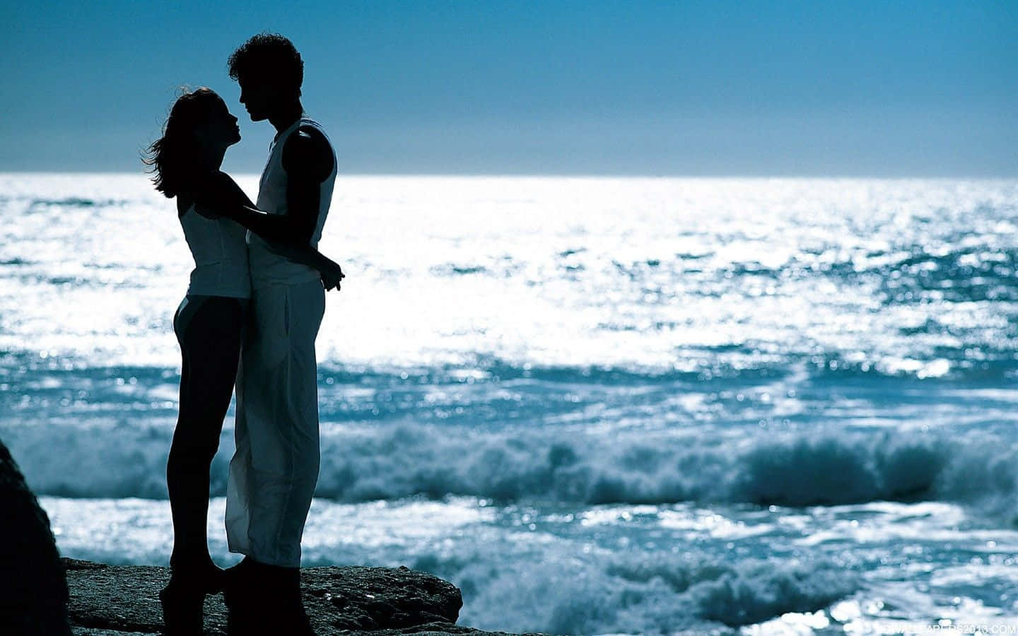 Beautiful Romantic 桌布 壁紙 — 一對剪影情侶在岩石海岸上擁抱，背後是陽光普照的海浪在清澈藍天下拍打。