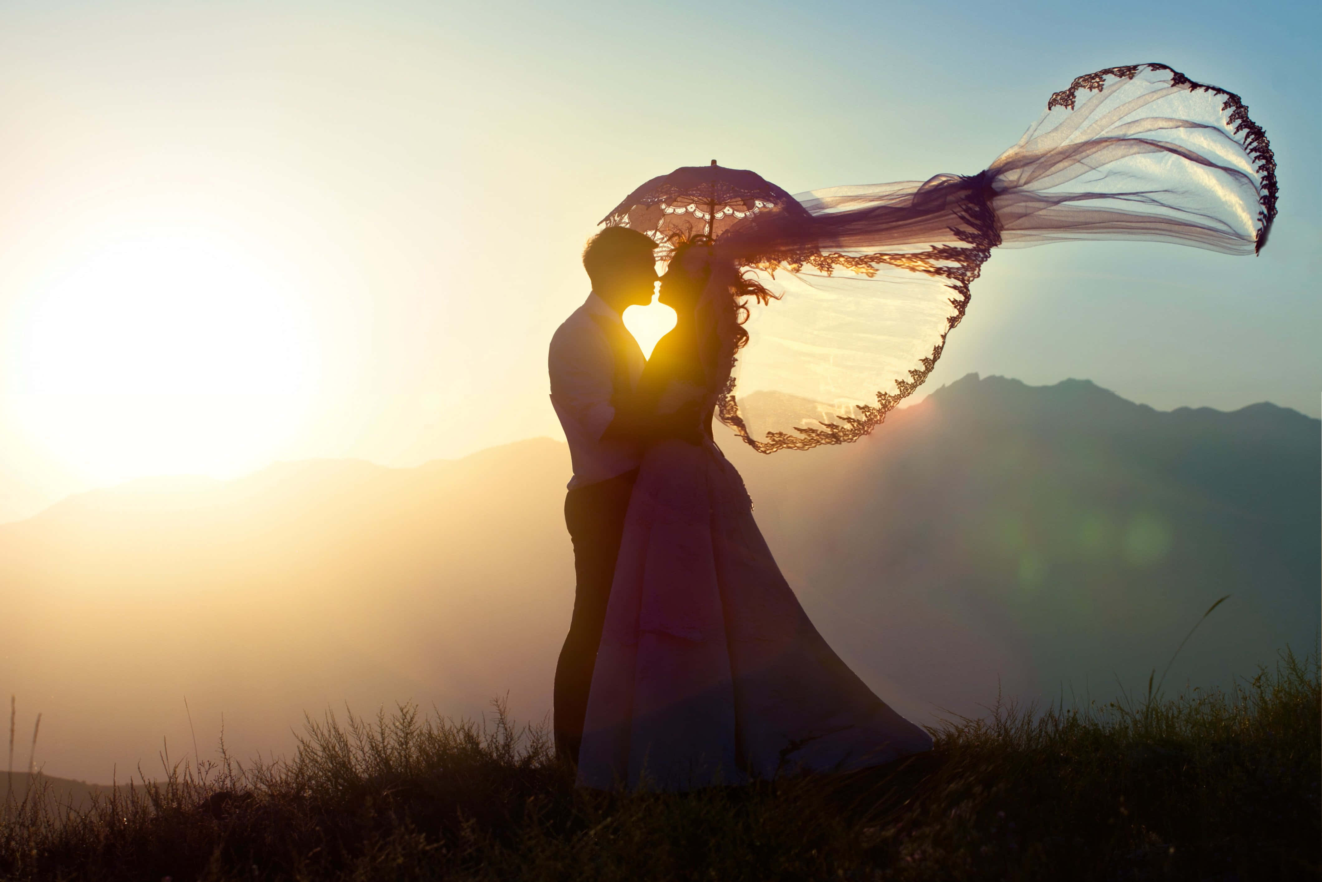 Wedding Couple 桌布 壁紙 — 一對剪影情侶在心形傘下接吻，背後山脊上金色夕陽光芒四射。